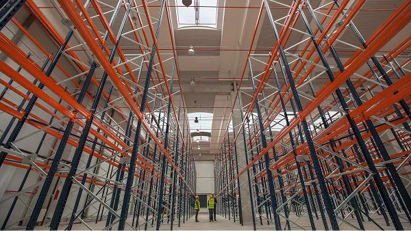 Interior of a warehouse with tall orange storage racking and a worker in a high-visibility vest.