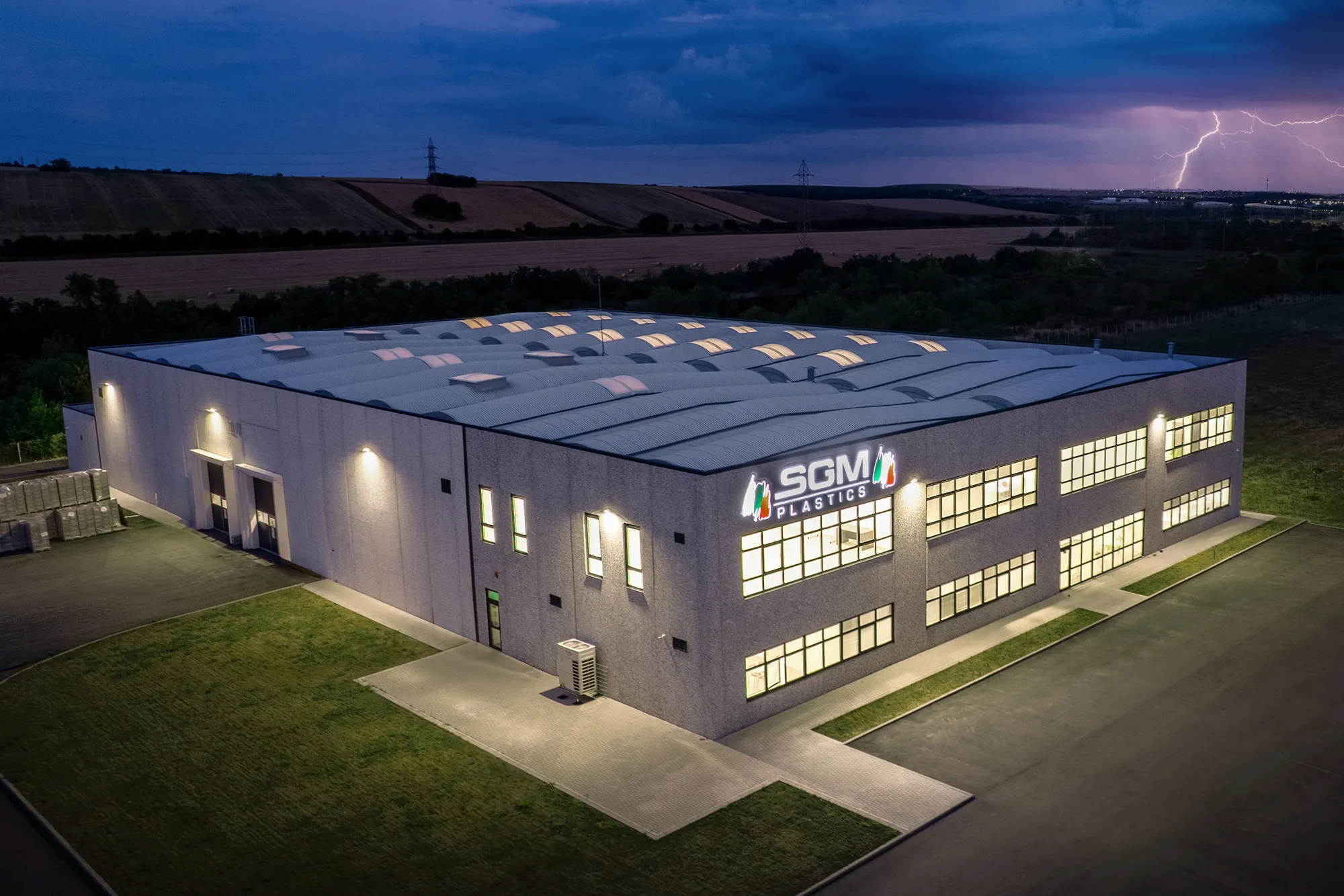 SGM Plastics factory building illuminated at dusk with a lightning storm in the distance.