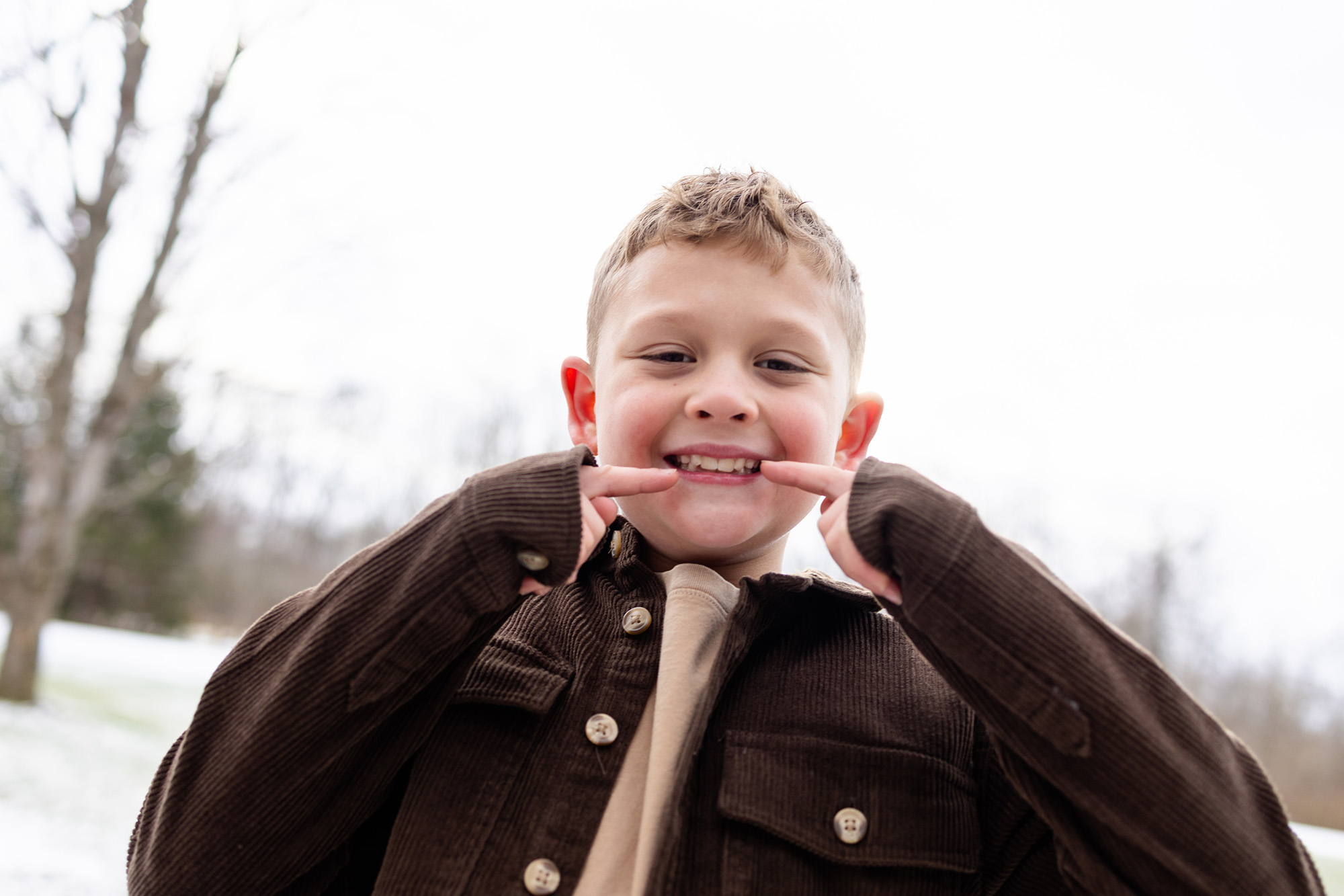 Smiling young boy outdoors pointing to his teeth wearing a brown jacket on a cloudy day.