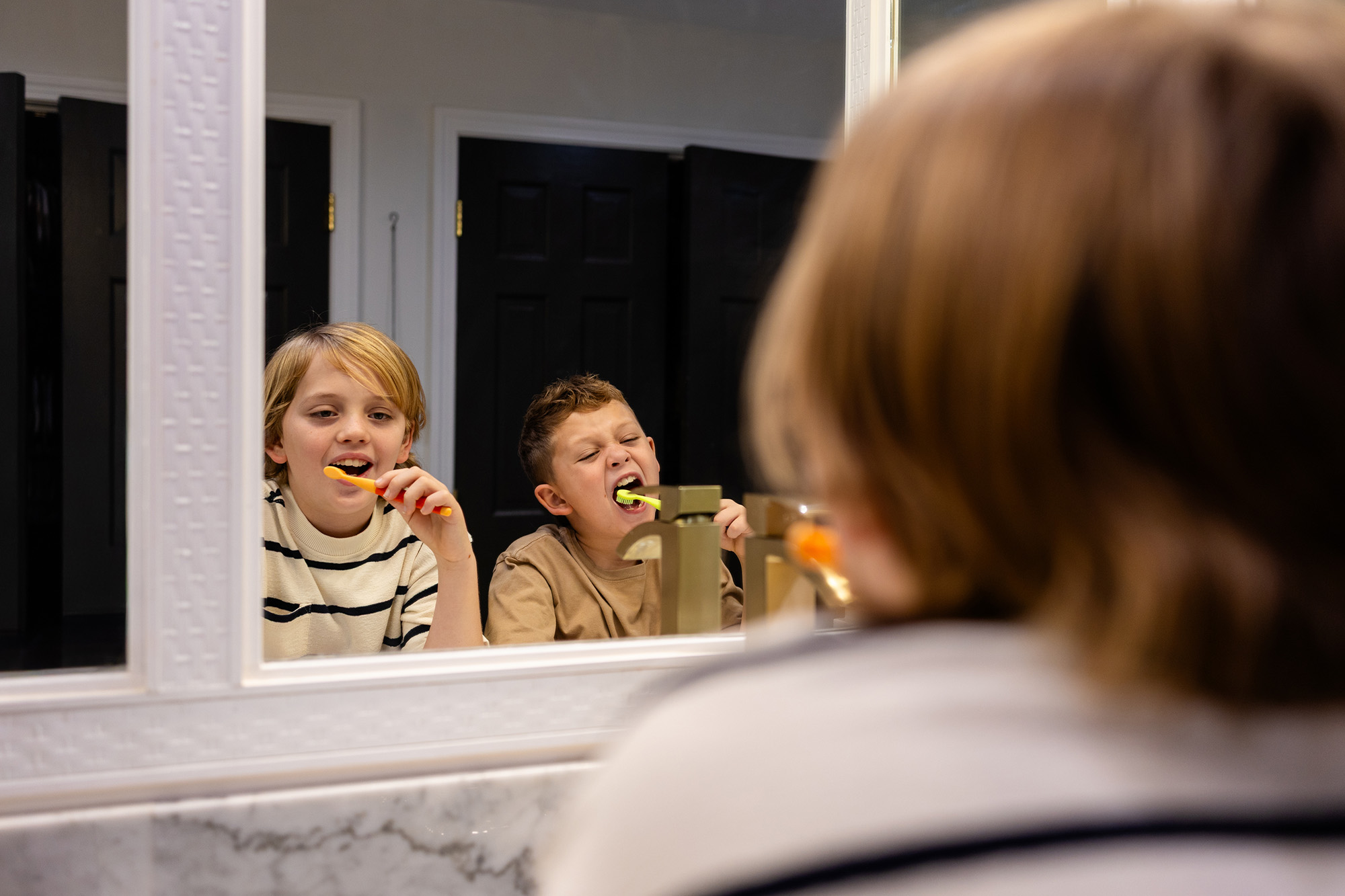 Two boys brushing their teeth in front of a bathroom mirror with black doors behind them.