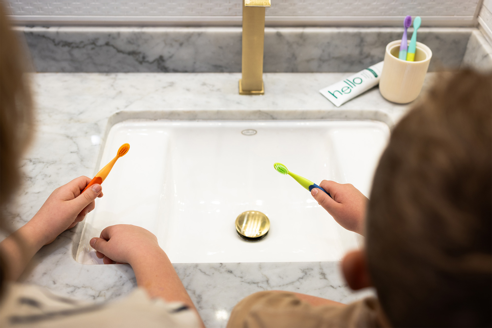Two children holding colorful toothbrushes over a white marble sink with a gold faucet.
