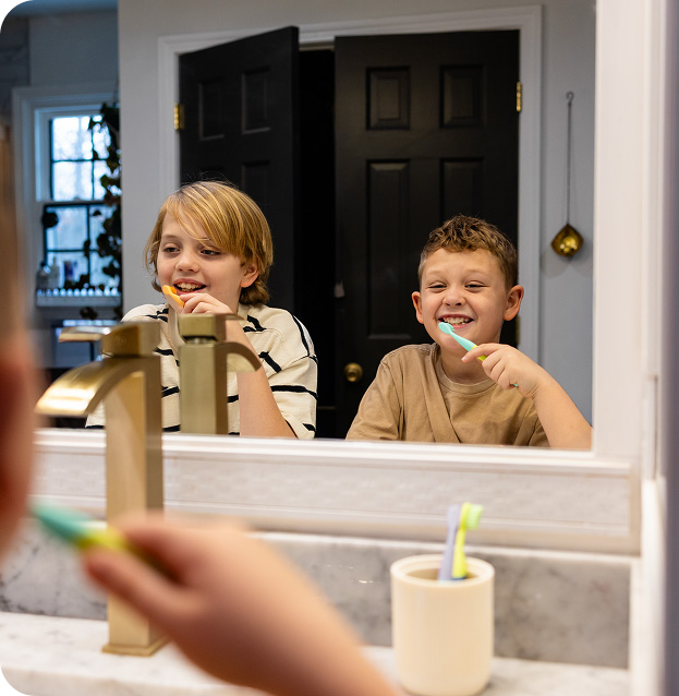 Two boys smiling while brushing their teeth in front of a bathroom mirror.