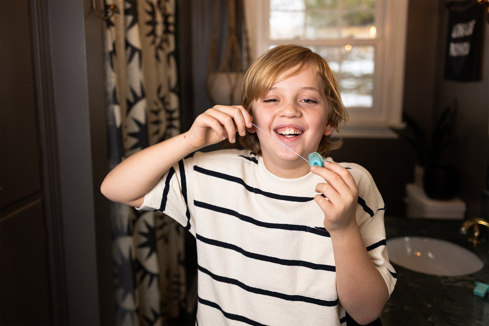 Smiling child in striped shirt flossing their teeth with a blue floss container in a bathroom.