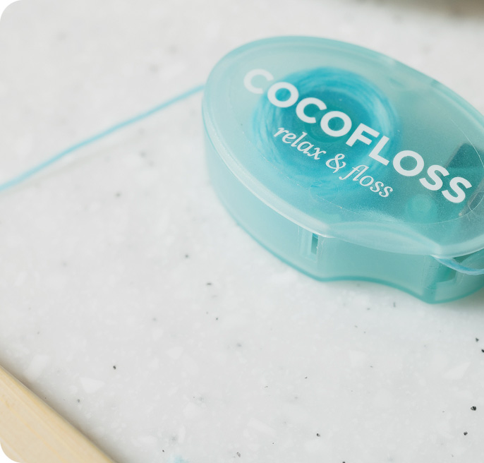 Light blue COCOFLOSS dental floss container on a white speckled surface with floss partly unwound.