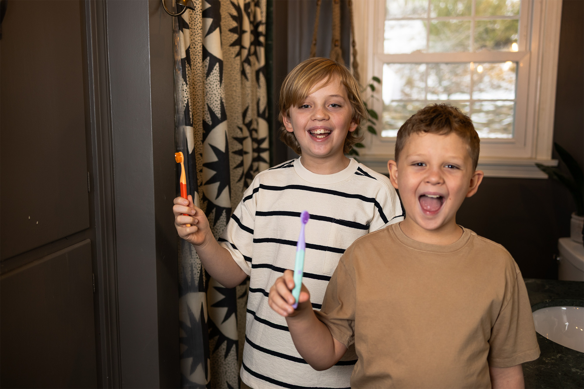 Two boys in a bathroom holding toothbrushes, smiling and laughing near a sink and window.