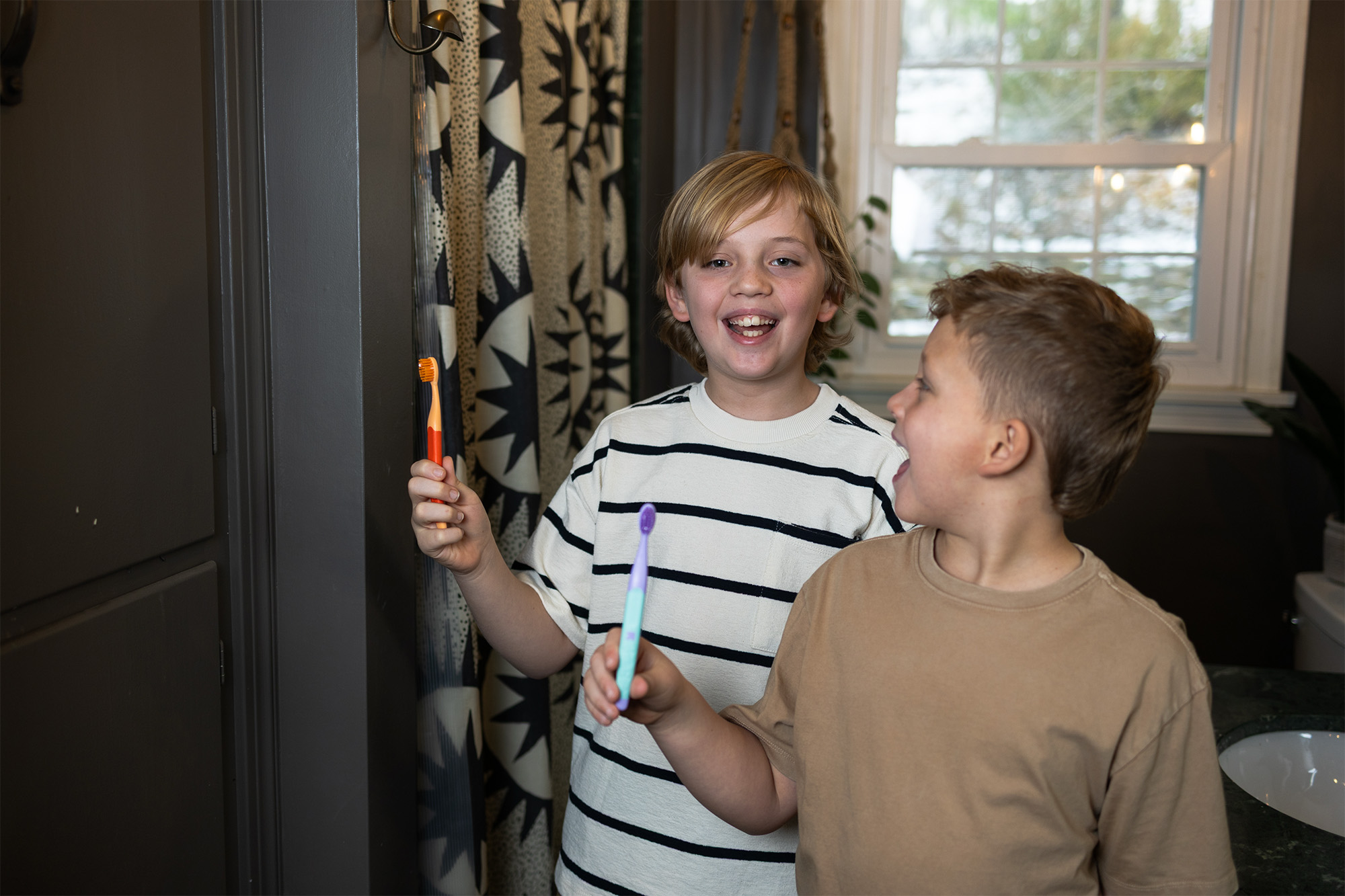 Two boys in bathroom holding toothbrushes and smiling, one wearing a striped shirt.