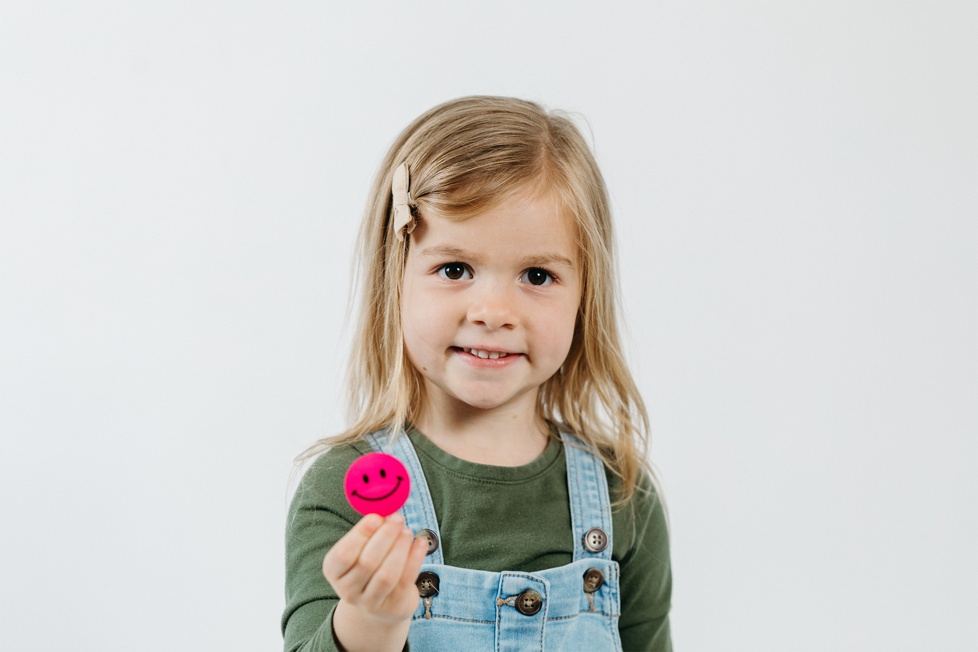 Young girl with blonde hair holding a pink smiley face token while smiling at the camera.