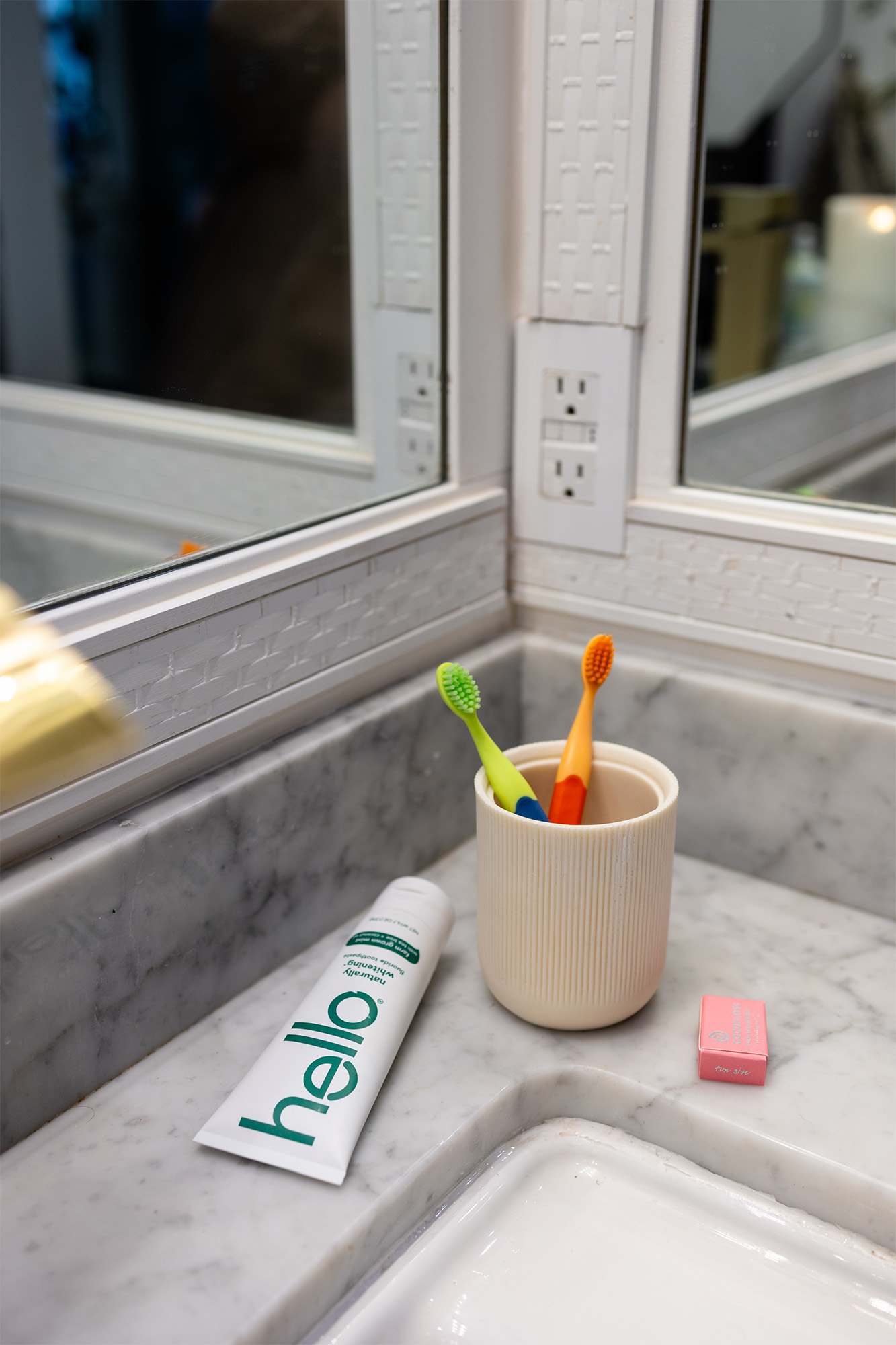 Bathroom countertop with two toothbrushes in a cup, a tube of Hello toothpaste, and a pink box of floss.