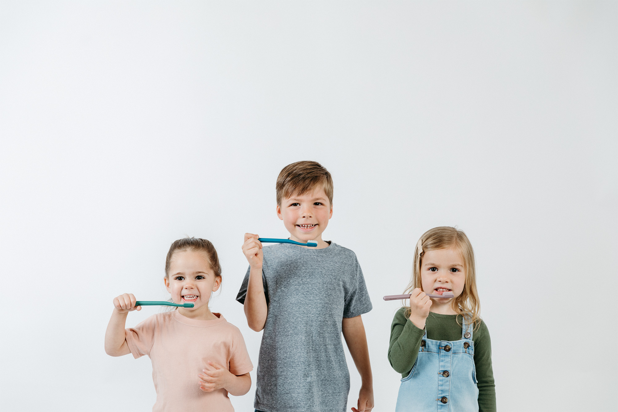 Three children smiling and brushing their teeth with colorful toothbrushes against a white background.