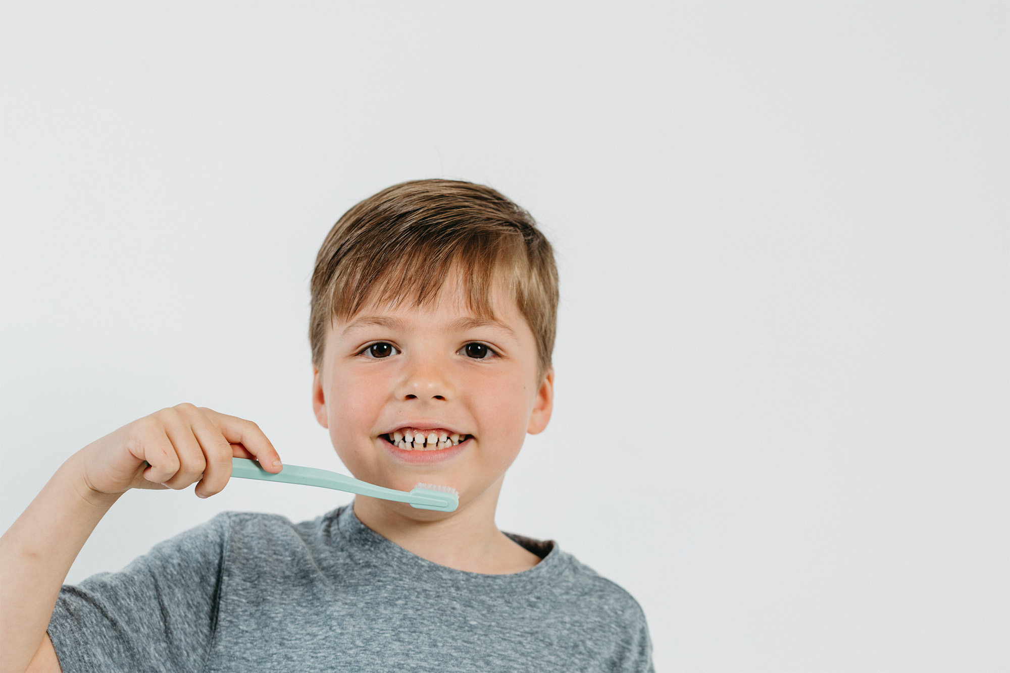 Smiling boy in gray shirt holding a mint green toothbrush near his teeth against white background.