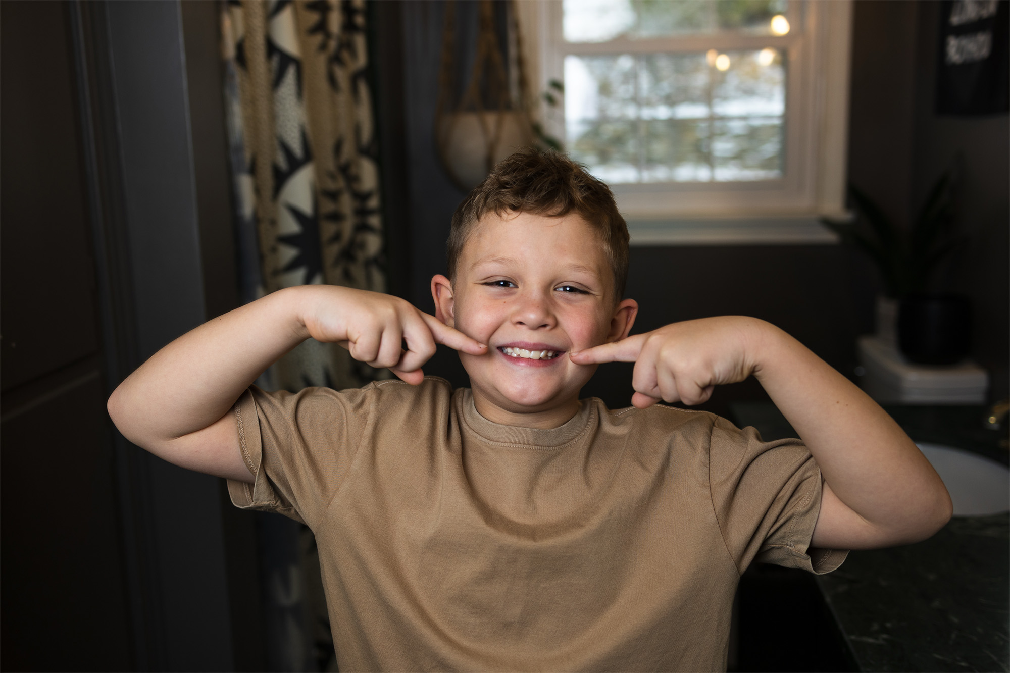 Smiling young boy in tan shirt pointing to his teeth in a home bathroom with window in background.
