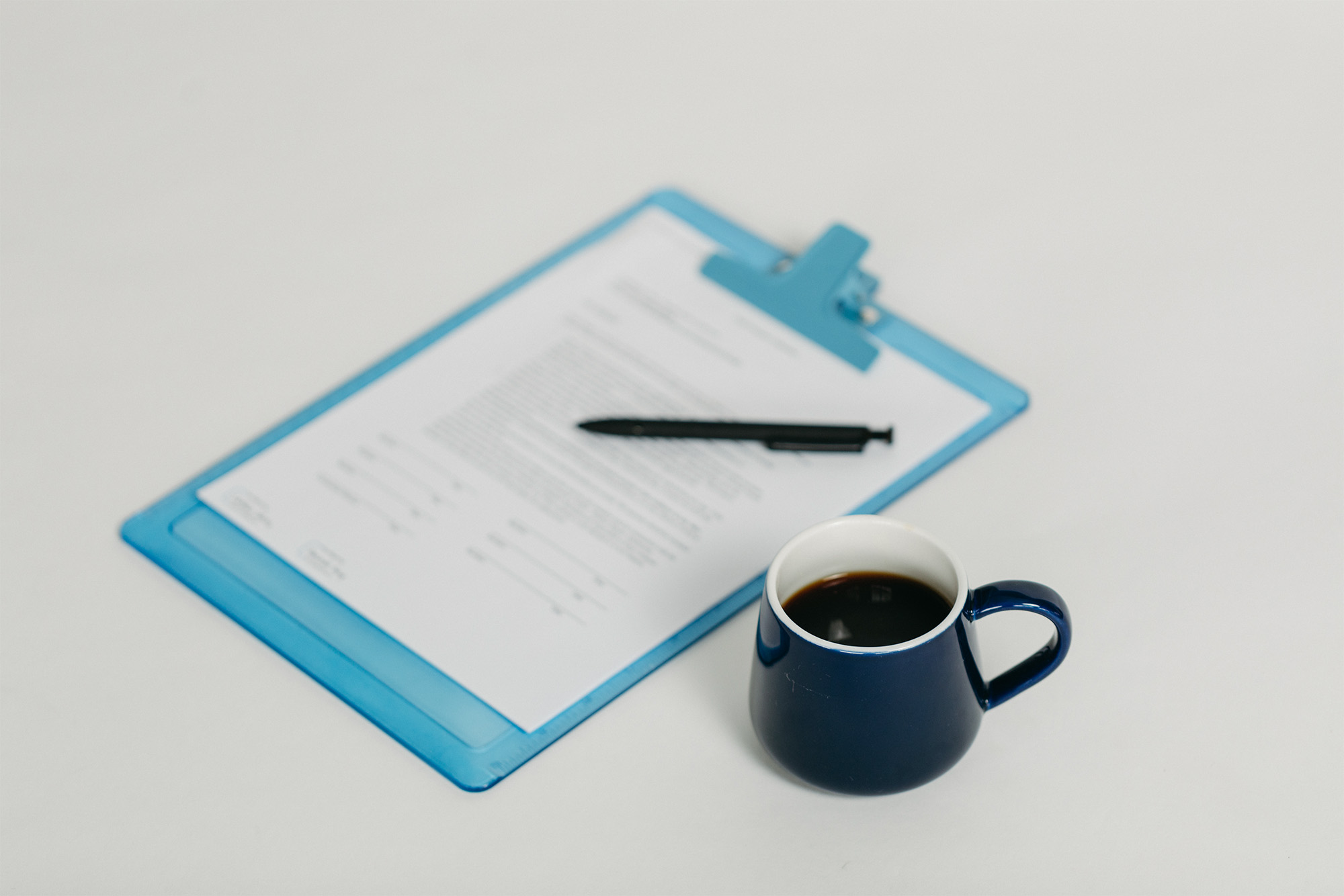 Blue clipboard with a document and pen beside a navy blue mug filled with coffee on a white surface.