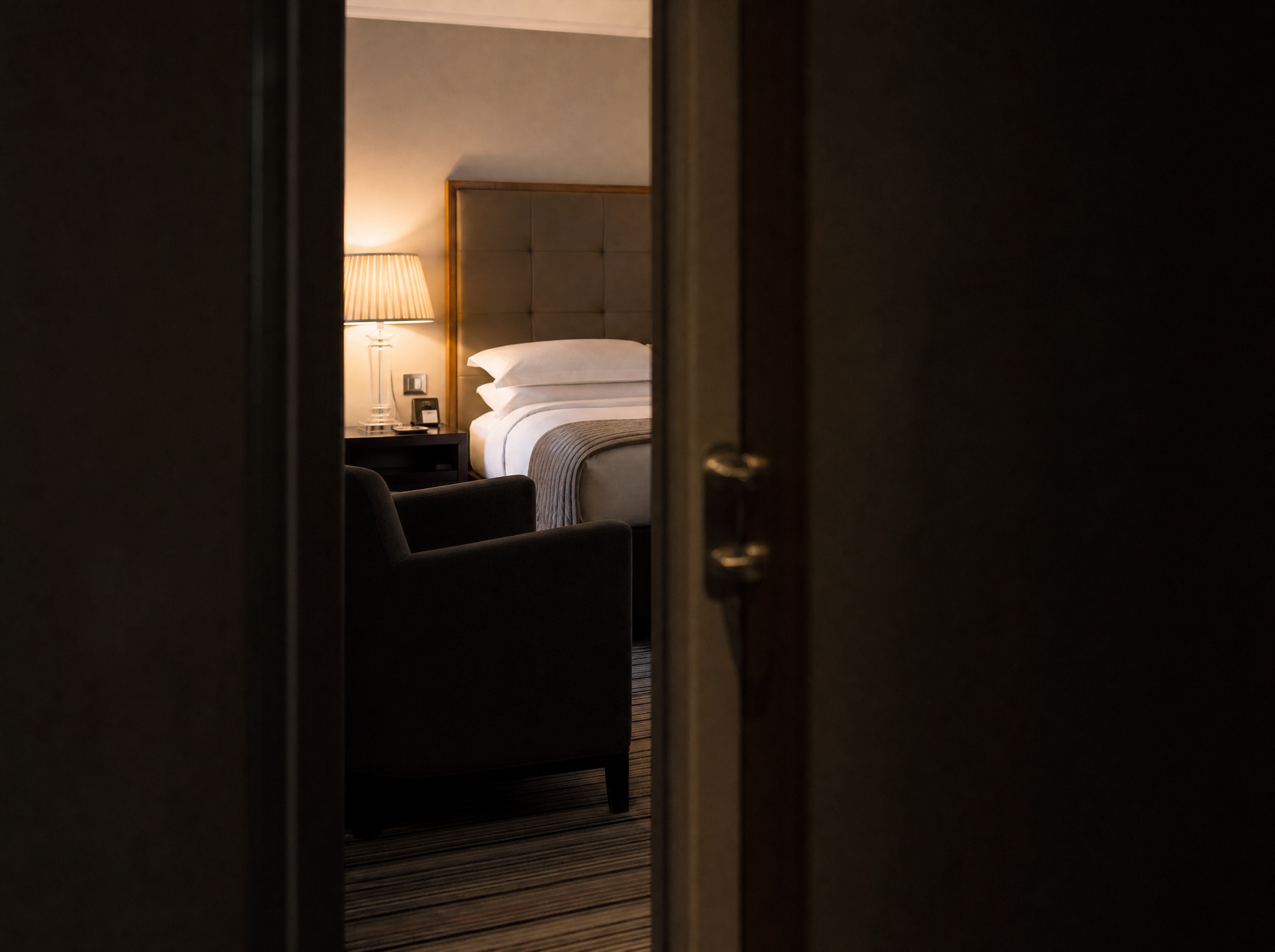 A hotel bedroom seen through a doorway with a bedside lamp lit