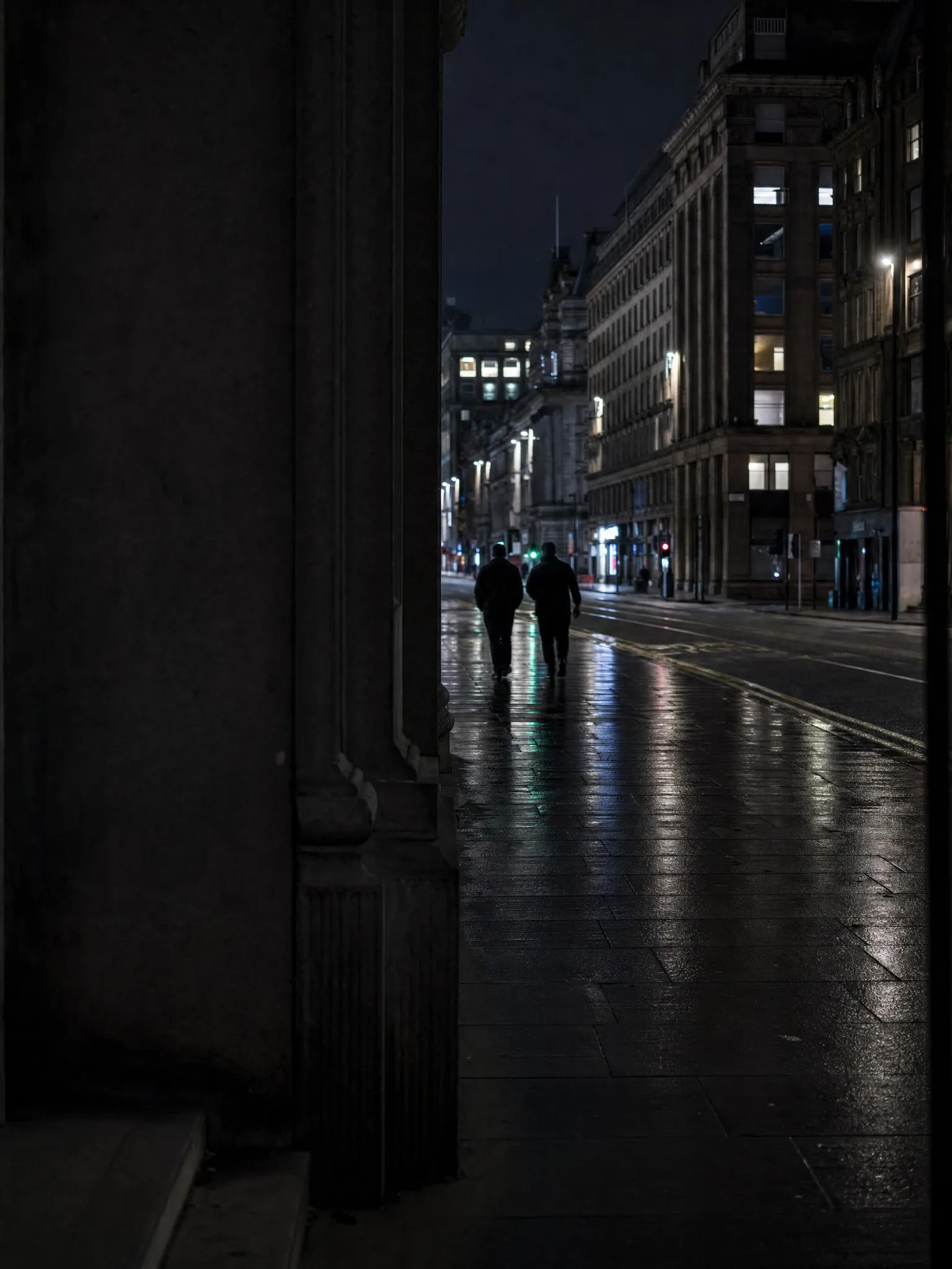 Two figures walking along a wet city centre street at night past lit office buildings