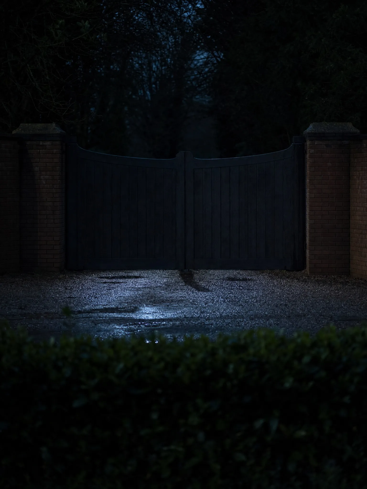 Closed timber gates between brick piers on a wet drive at night