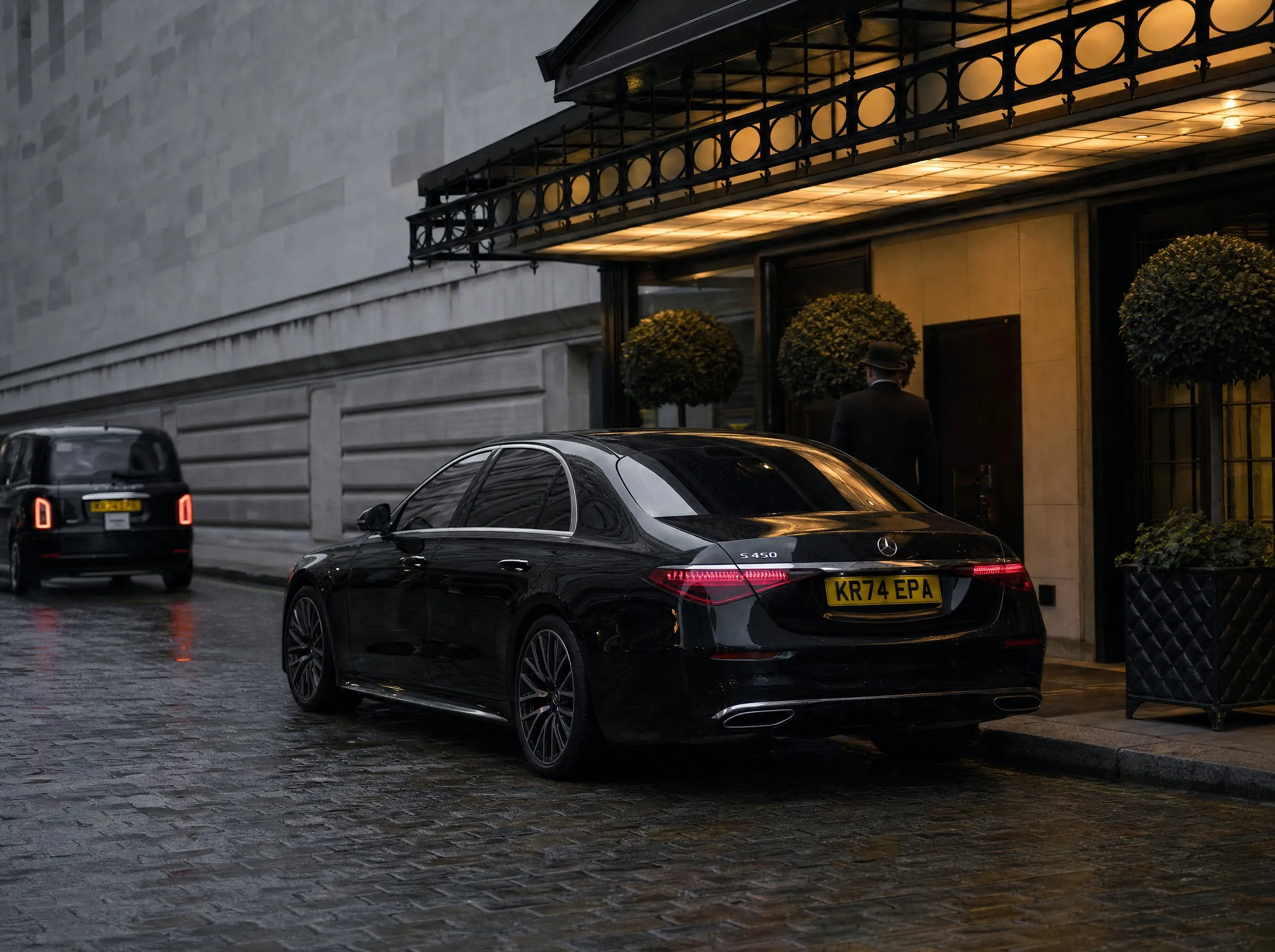 A lit hotel canopy at night with a doorman and a car waiting at the kerb