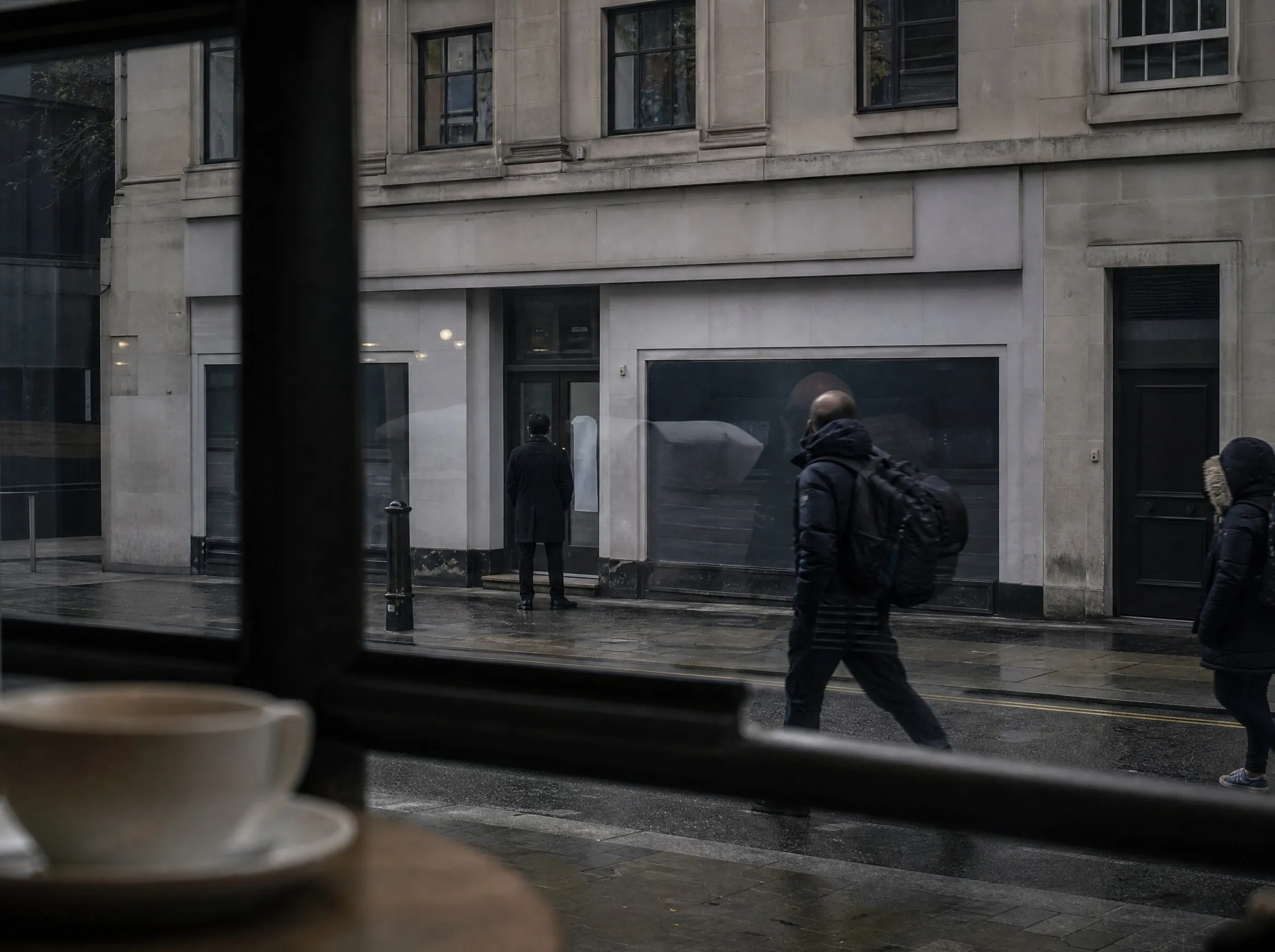 A wet street seen through a cafe window, a coffee cup in the foreground and people passing