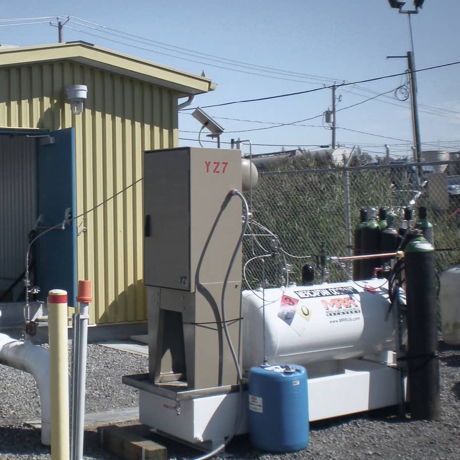 Small tan modular gas odorization building connected to storage tanks and piping at a rural renewable natural gas site, with utility poles in the background.