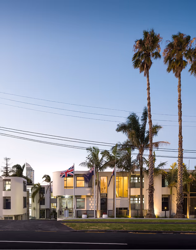 Modern two-story building with palm trees and flagpoles displaying British and other flags against a clear evening sky.
