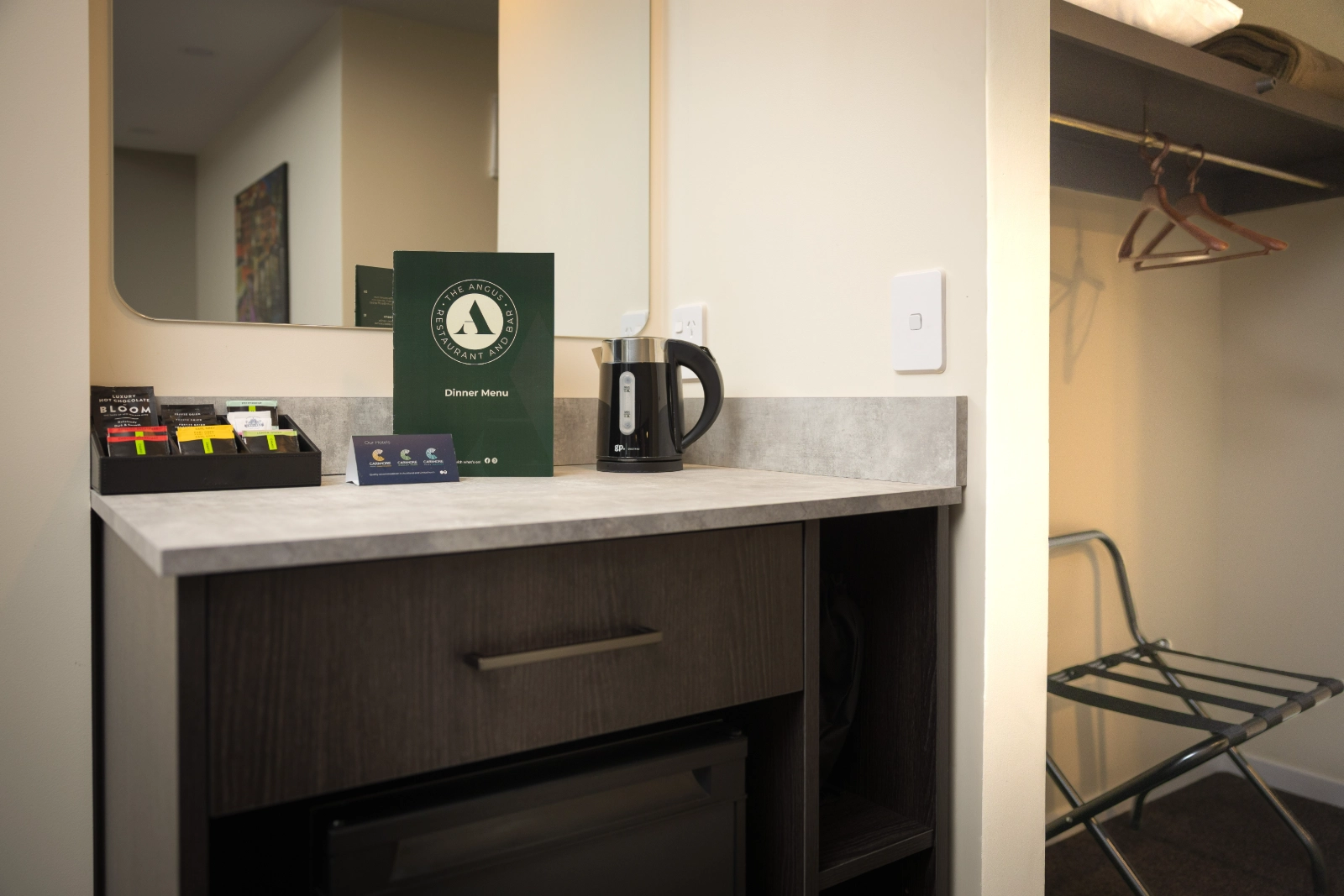 Hotel room counter with kettle, menu, tea packets, mirror, and open closet with hangers and luggage rack.