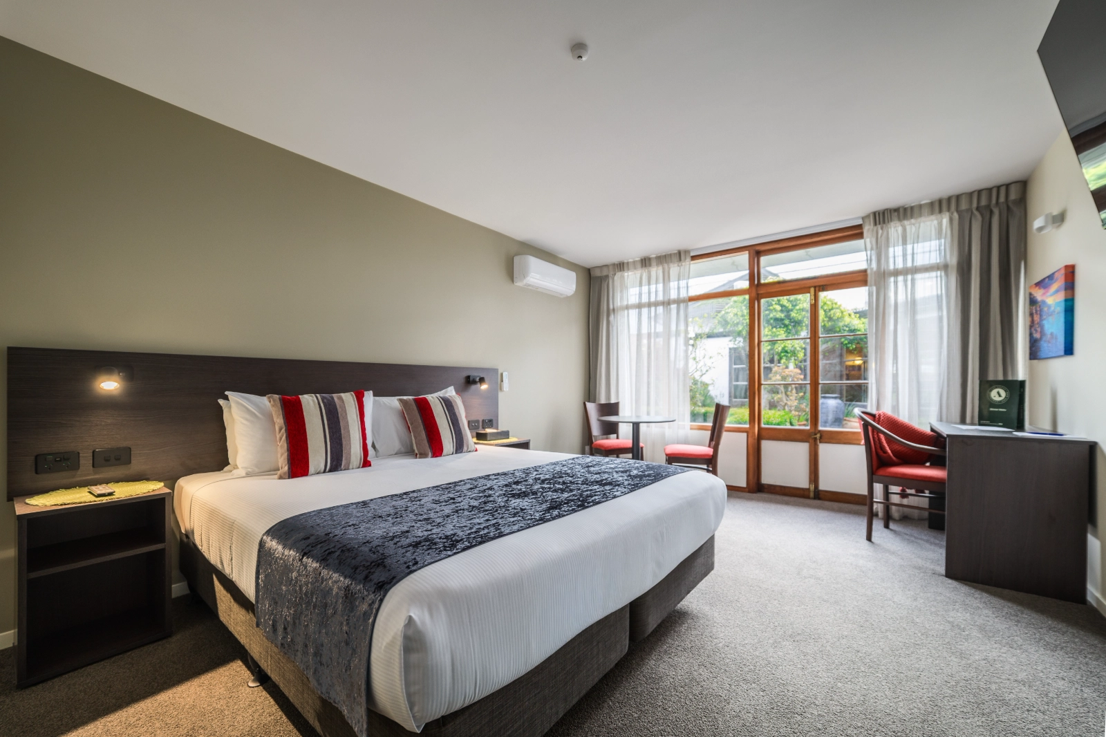 Hotel room with a king bed, striped pillows, a small table with chairs, and a window with garden view.