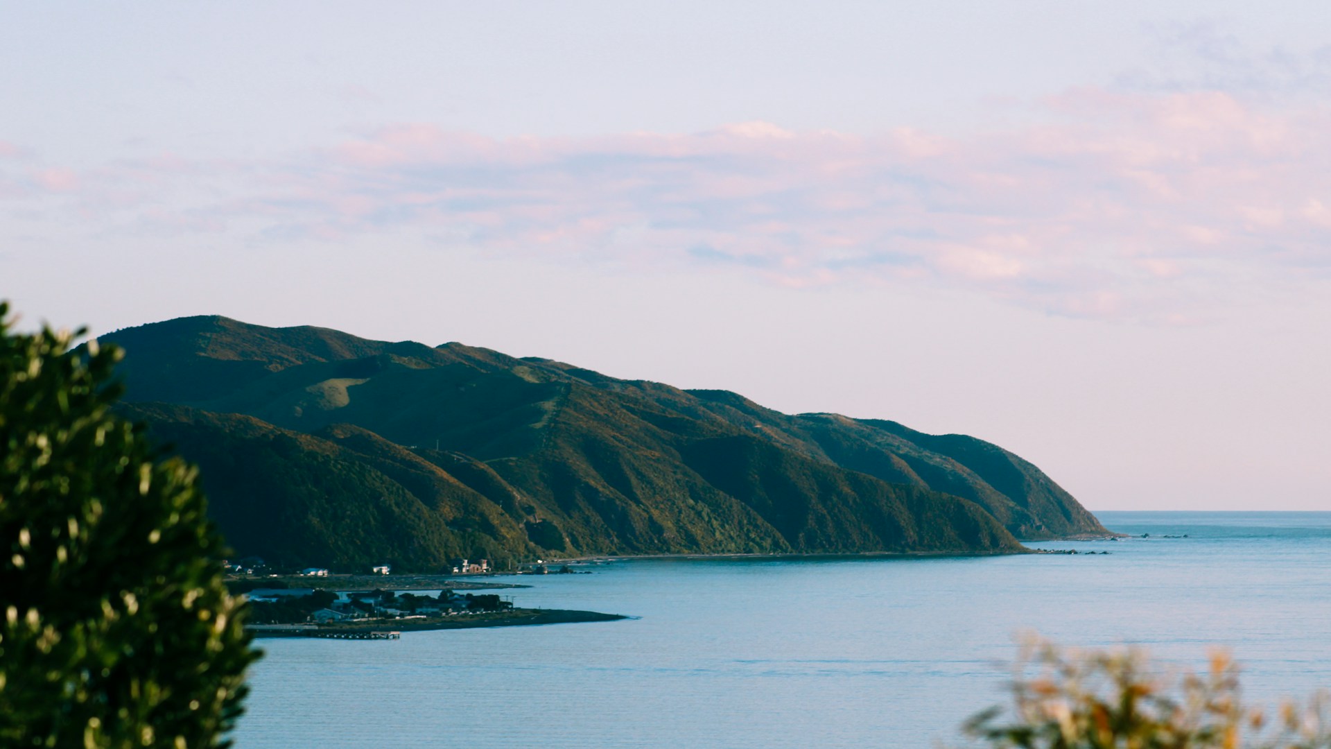 Coastal landscape with green hills, calm blue water, and a clear sky with scattered clouds.