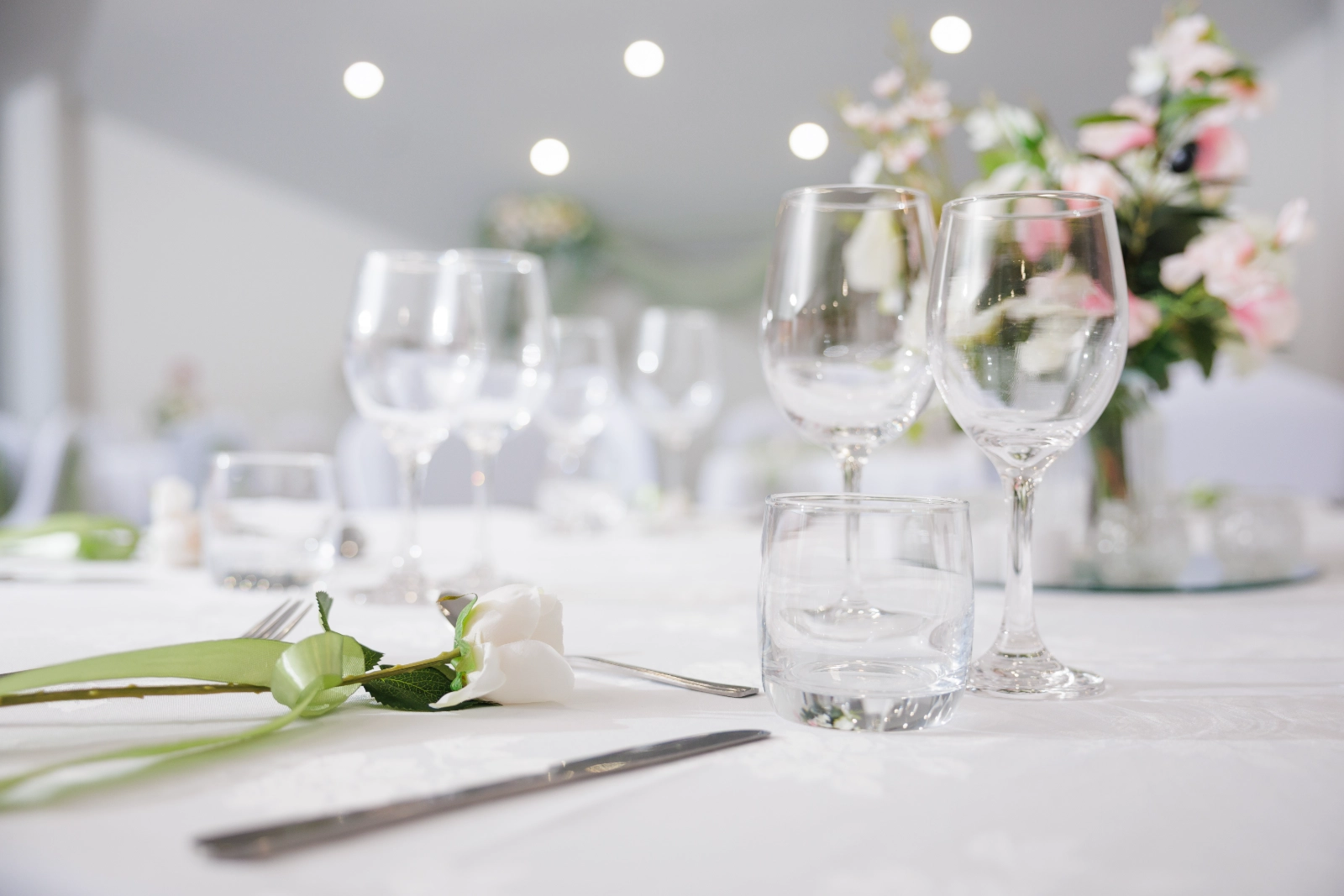 Elegant table setting with crystal glasses, silverware, and white rose on a white tablecloth.
