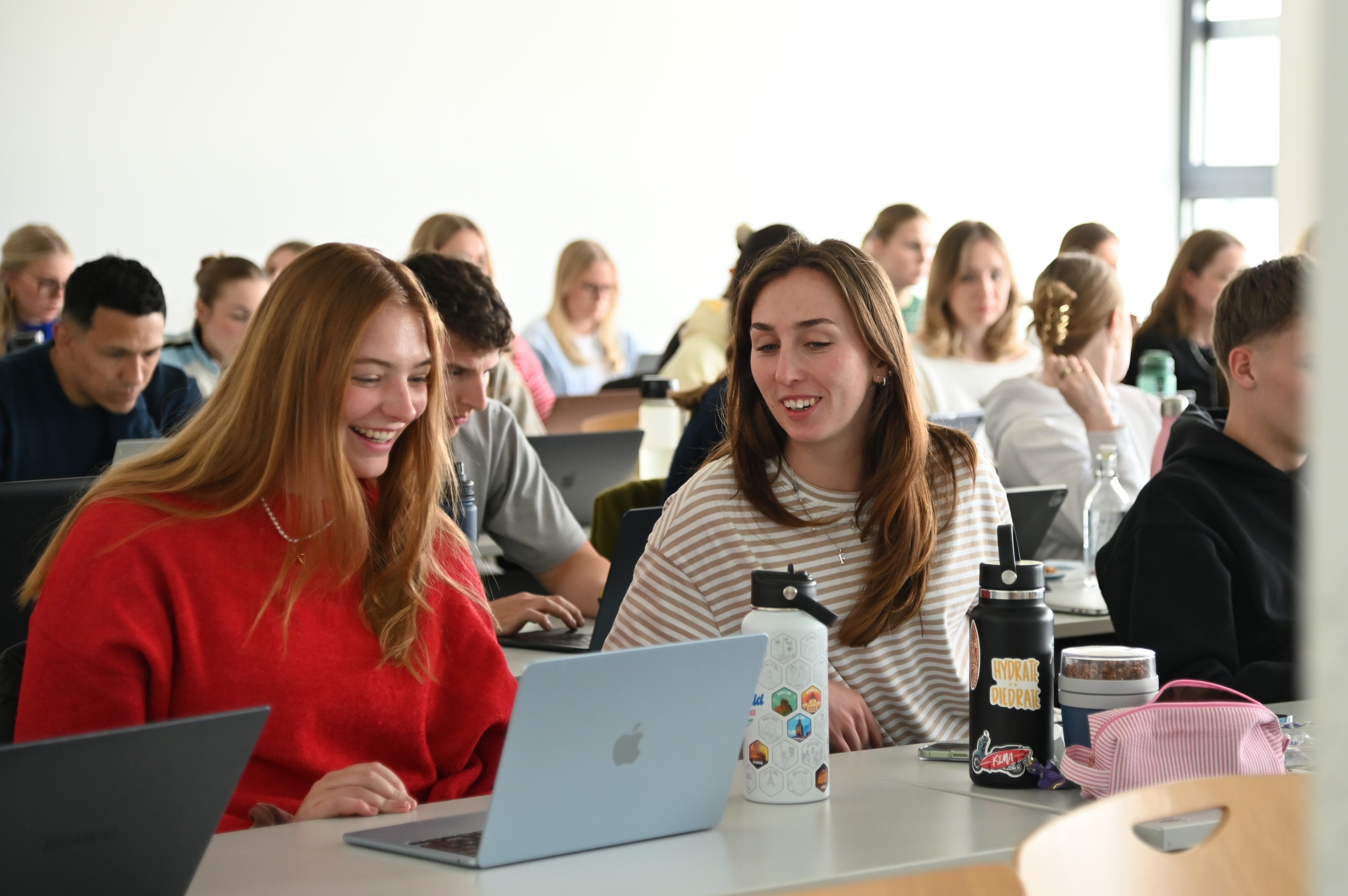 Students sitting in a classroom, two girls smiling and looking at a laptop together.
