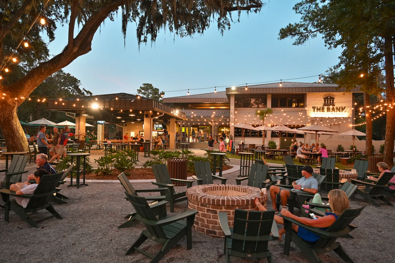 Outdoor seating area with string lights, fire pits, and people at tables near The Bank restaurant at dusk.