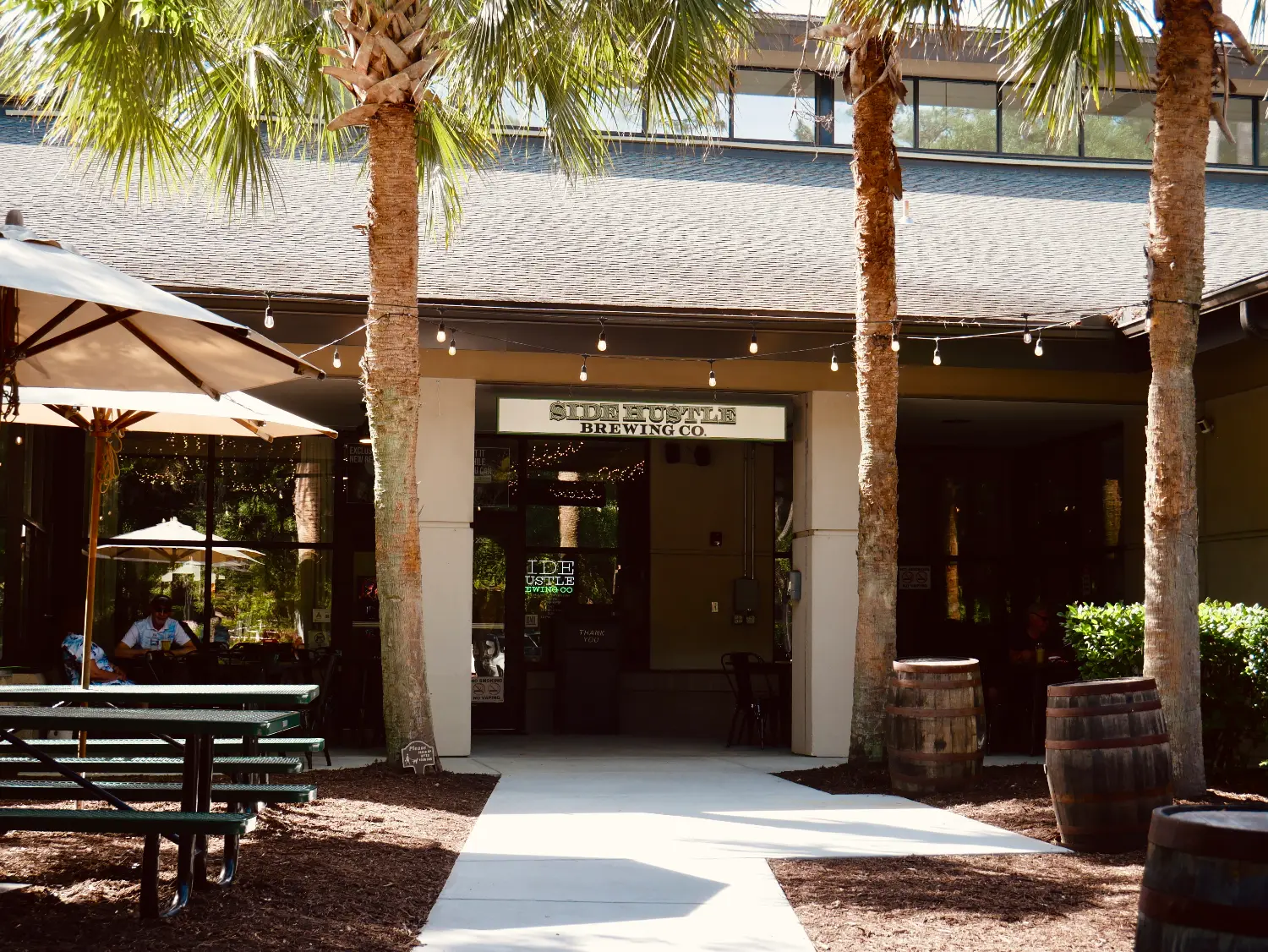 Outdoor entrance of Side Hustle Brewing Co. with palm trees, picnic tables, and barrels.