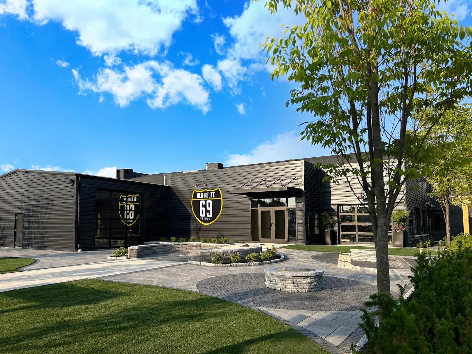 Modern black building with Old Route 69 Brewery sign, landscaped yard, trees, and blue sky with clouds.