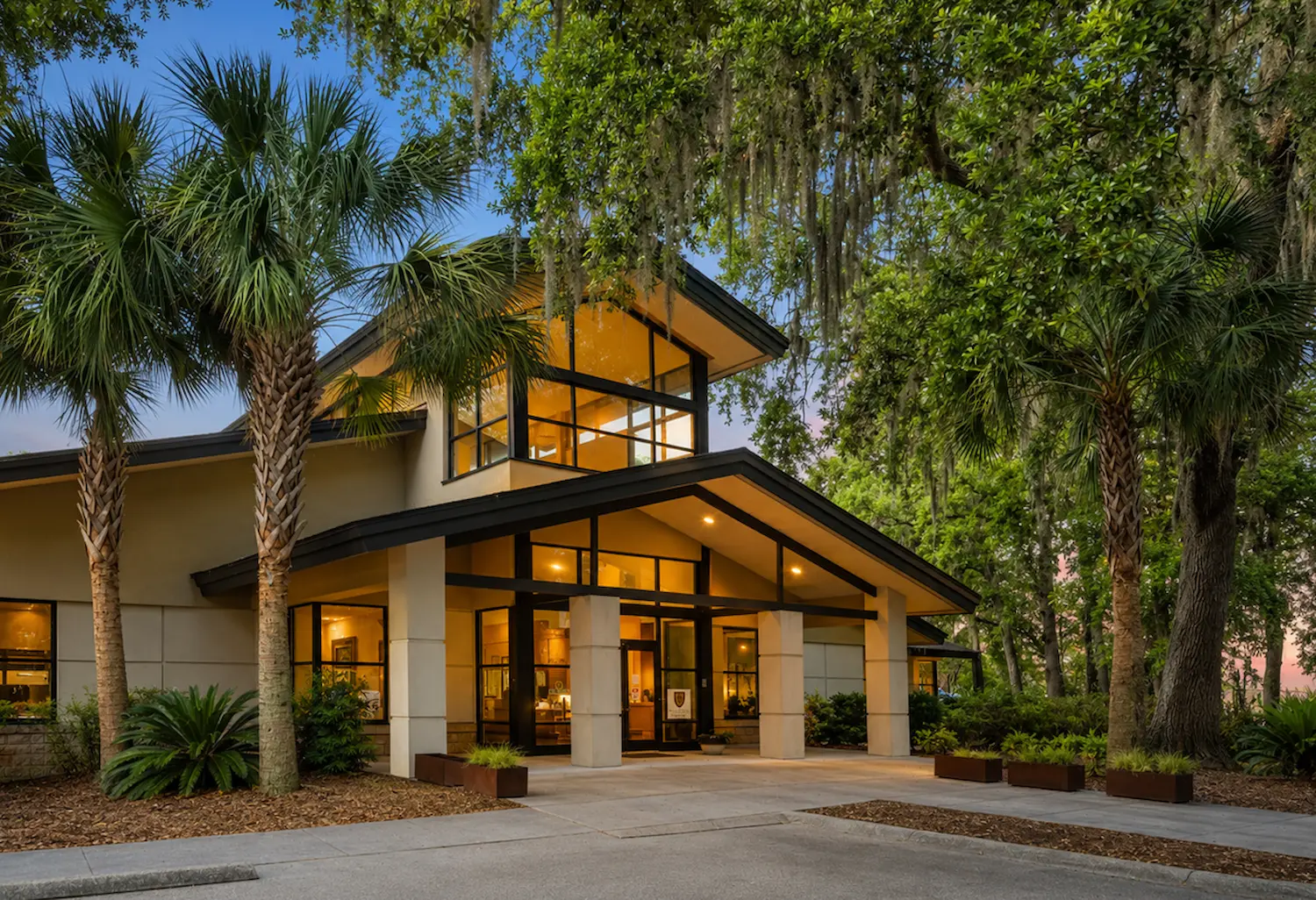 Modern building entrance with large glass windows, palm trees, and greenery at dusk.