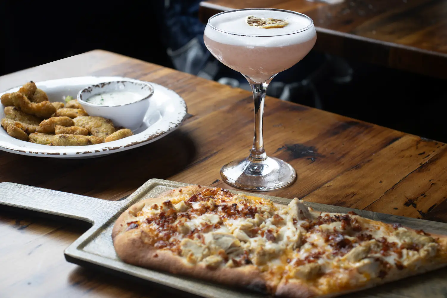 Chicken and bacon flatbread on a wooden board with a coupe cocktail and a plate of fried pickles with ranch on a dark wood table.