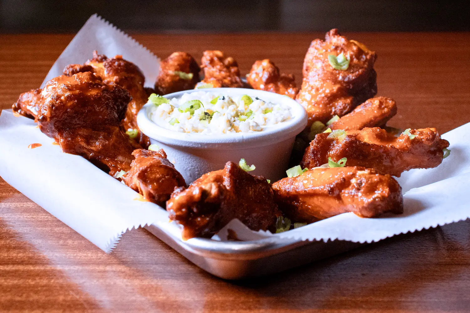 Basket of buffalo wings in a paper-lined basket with blue cheese dip and scallions, on a wood table.