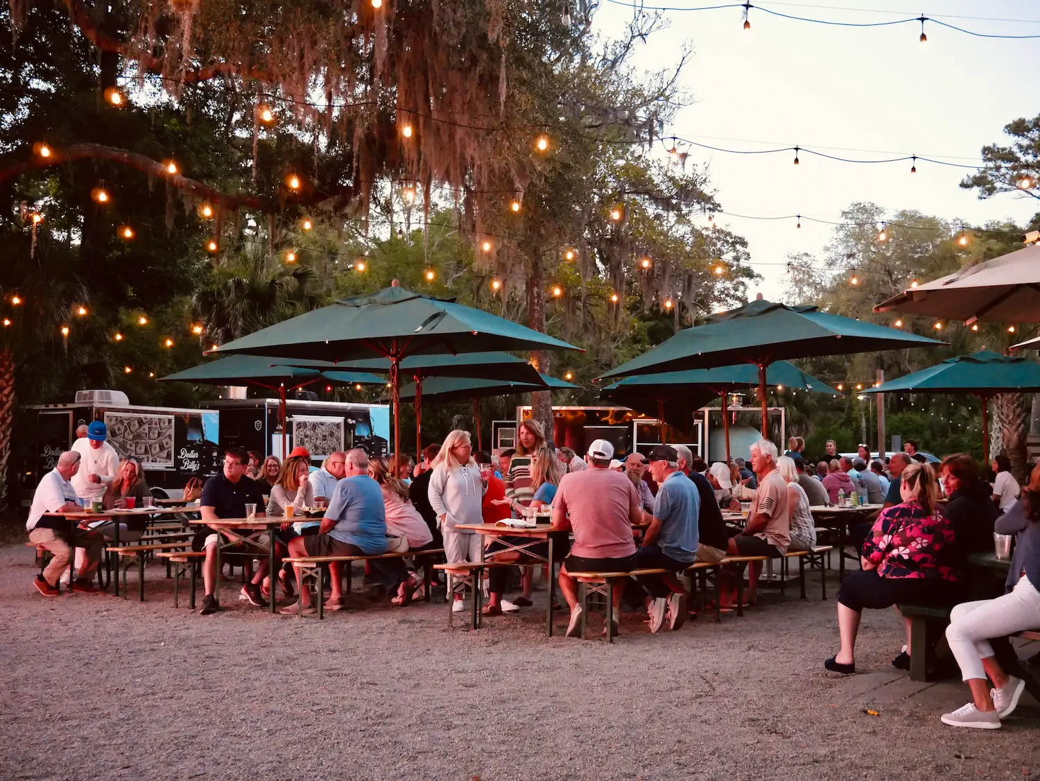 A crowd at communal picnic tables under string lights and umbrellas at dusk, with food trucks parked beneath moss-draped live oaks.
