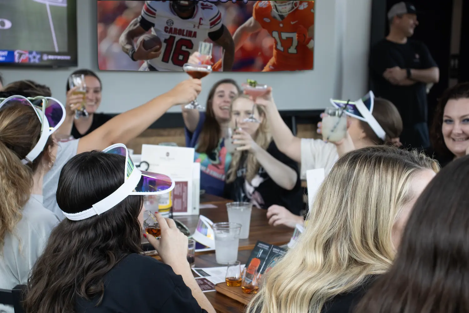 A bachelorette group in matching visors raising cocktails in a toast around a table, with TVs showing a football game behind them.