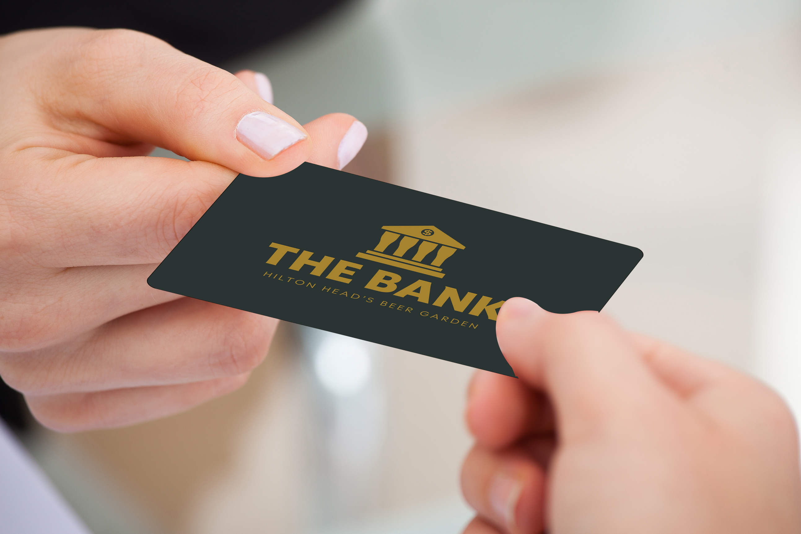 A gift card for The Bank, Hilton Head's beer garden, held between two hands with the gold columned logo.