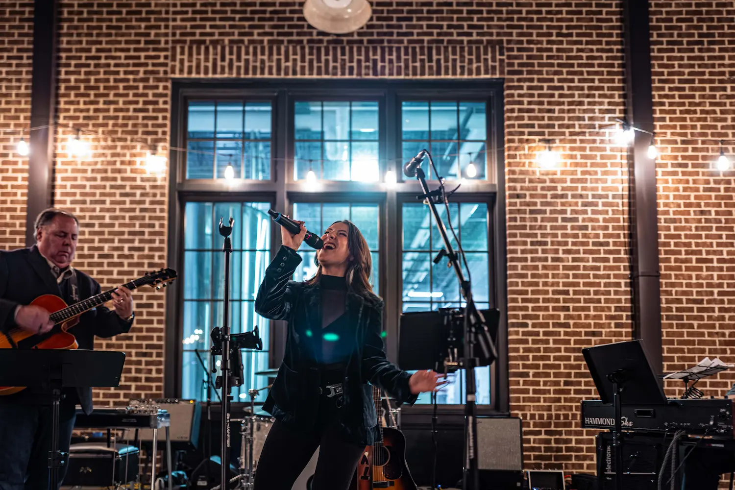 Woman passionately singing with microphone beside man playing guitar in a brick-walled live music venue.