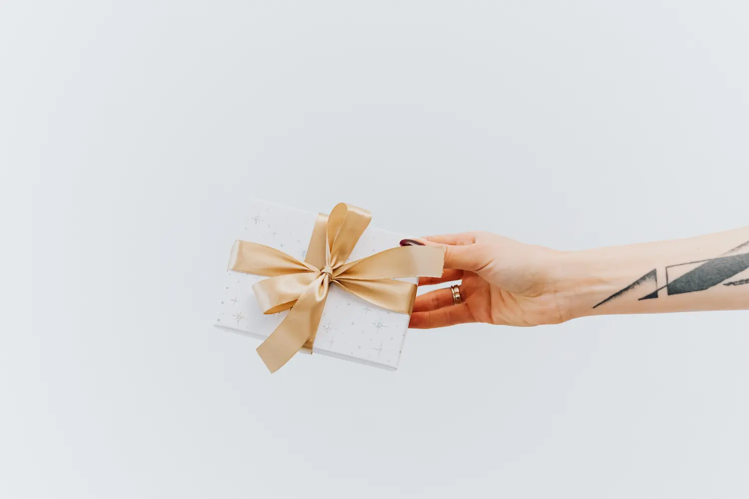 Hand with tattoo and rings holding a white gift box tied with a gold ribbon bow.