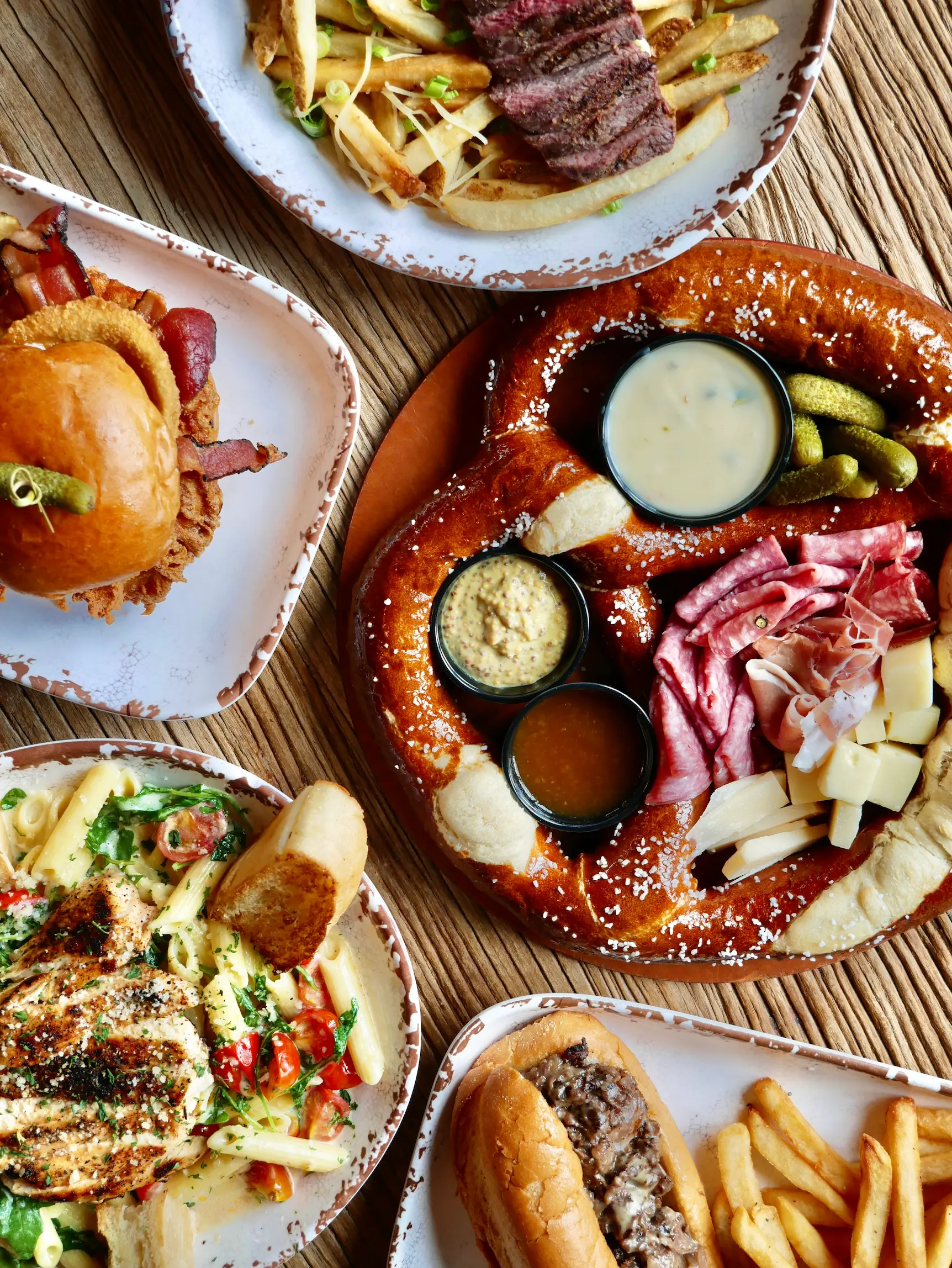 Overhead view of various dishes: pretzel with meats and sauces, sandwiches, fries, and grilled chicken pasta.
