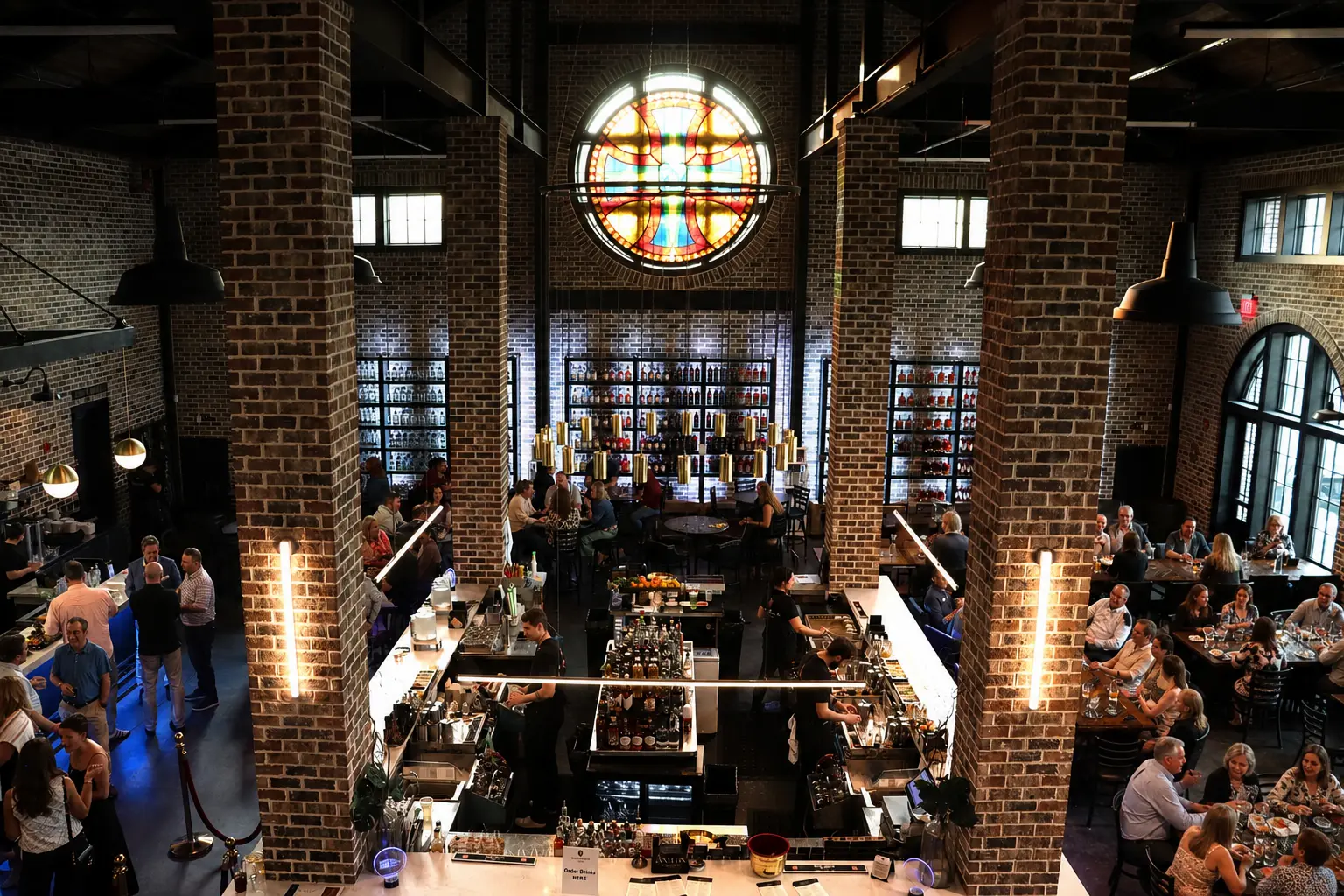 Interior of Burnt Church Distillery from above, with a stained glass rose window over the bar, exposed brick columns, and guests at tables below.