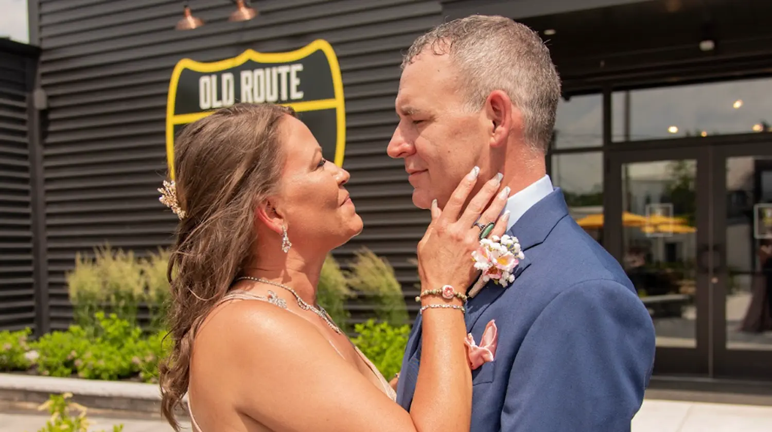 A couple in wedding attire embracing outside Old Route 69 Brewery, with the yellow shield logo on the building behind them.