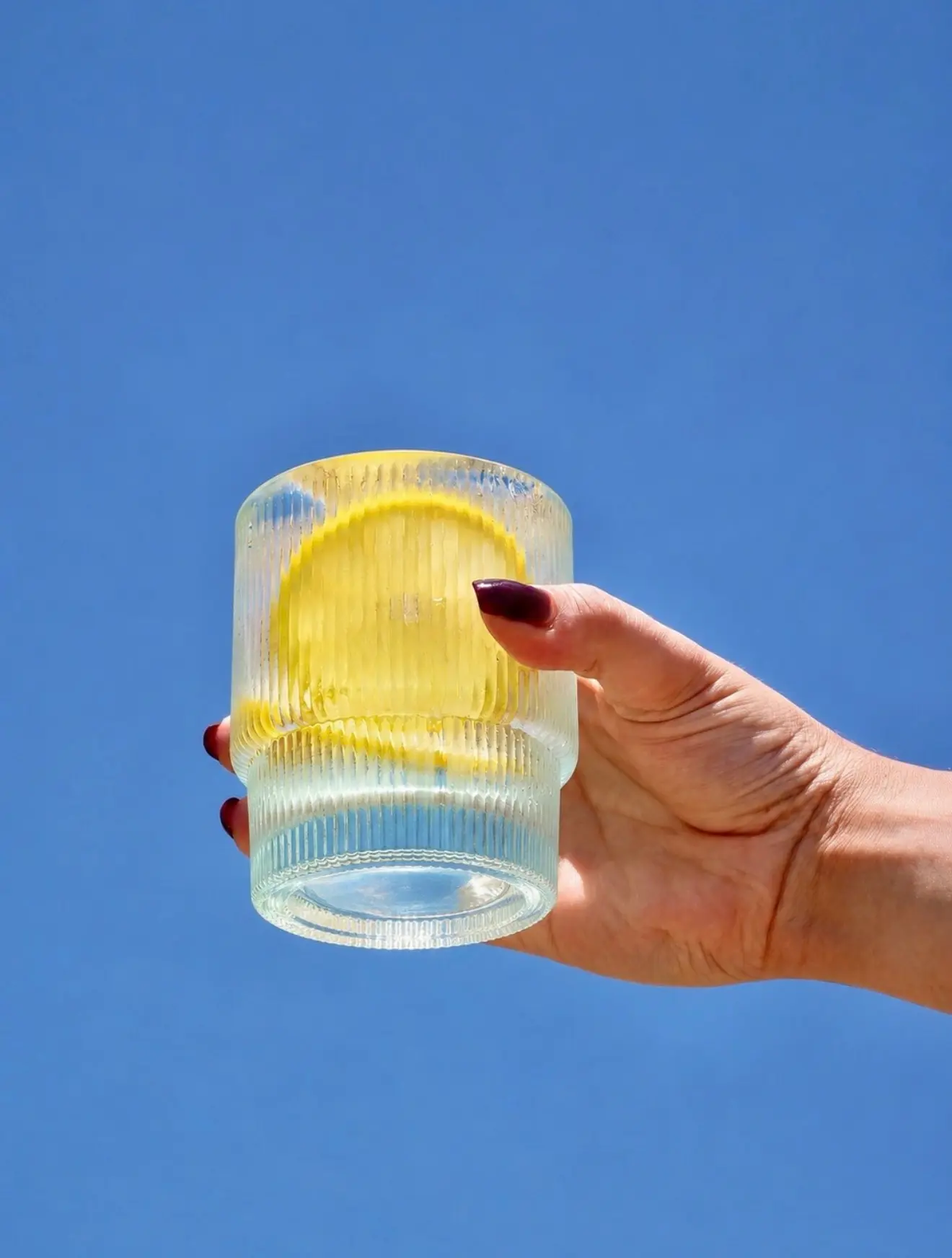 A hand holding up a ribbed glass tumbler with a clear drink and a lemon wheel against a bright blue sky.