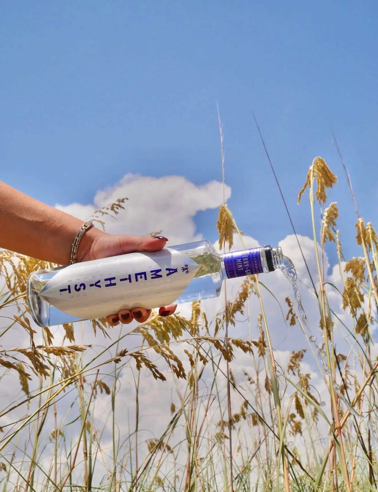 Hand pouring liquid from a clear bottle labeled Amethyst Berry Gin Mint in a grassy field under blue sky.