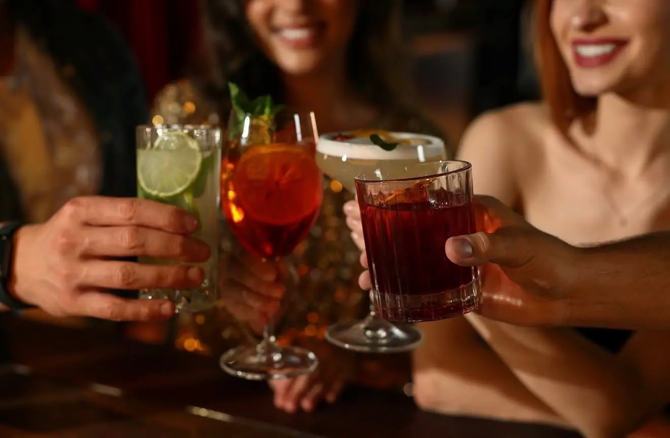 Four cocktails raised in a toast at a bar — a mojito, a spritz with orange, a foam-topped sour, and a dark drink on the rocks.