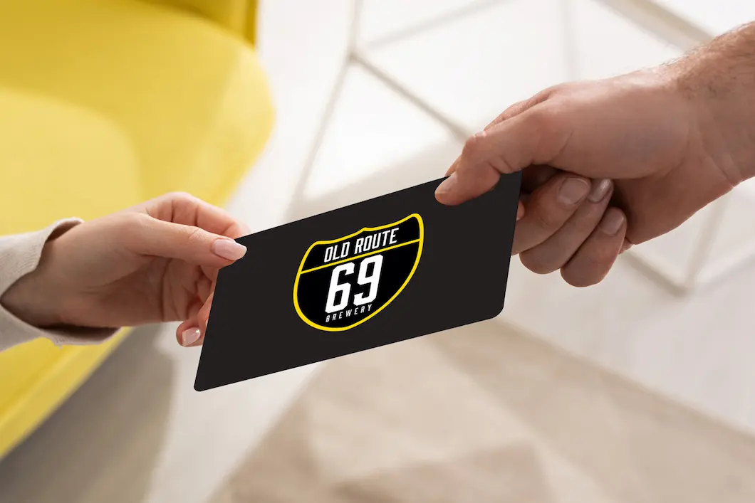 An Old Route 69 Brewery gift card being handed from one person to another, showing the yellow shield logo.