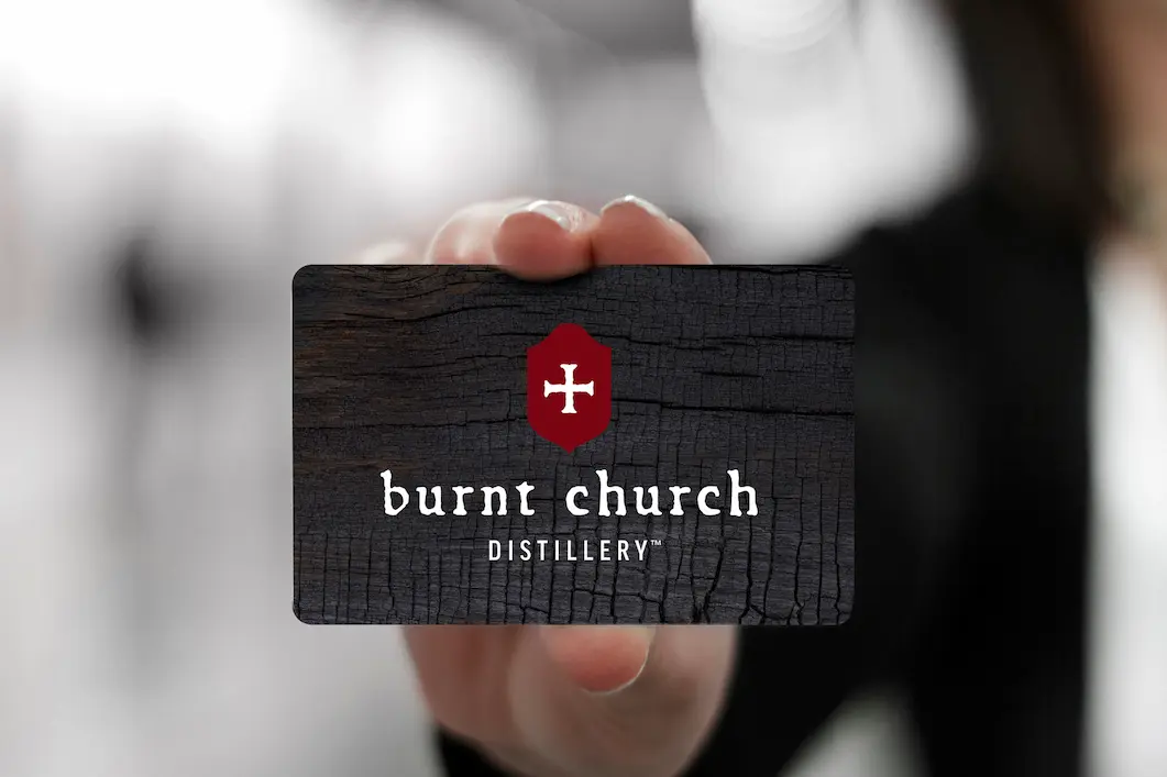 A hand holding a Burnt Church Distillery gift card with a charred wood design and the red cross logo.