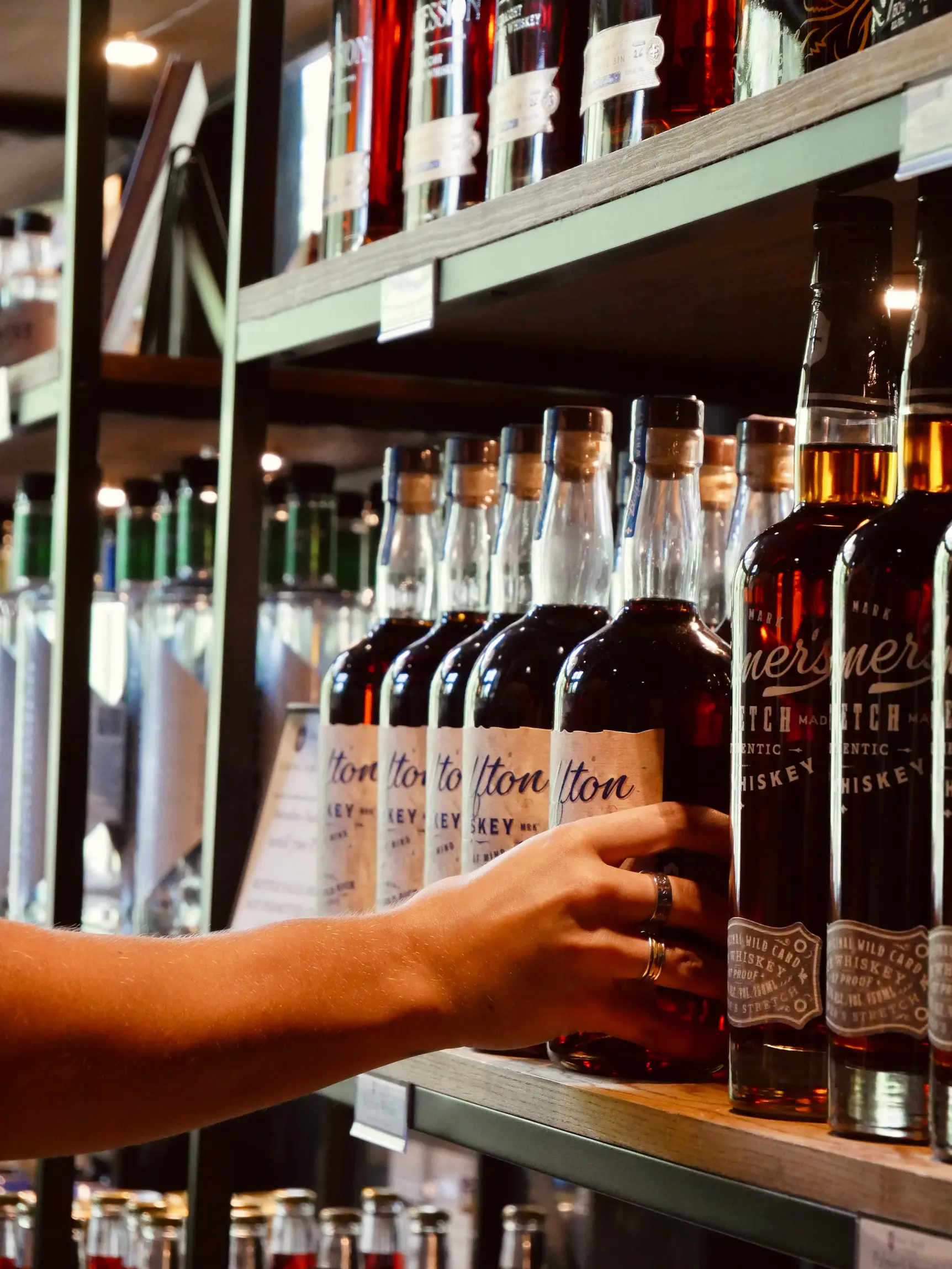Hand reaching for a whiskey bottle on a store shelf among rows of liquor bottles.