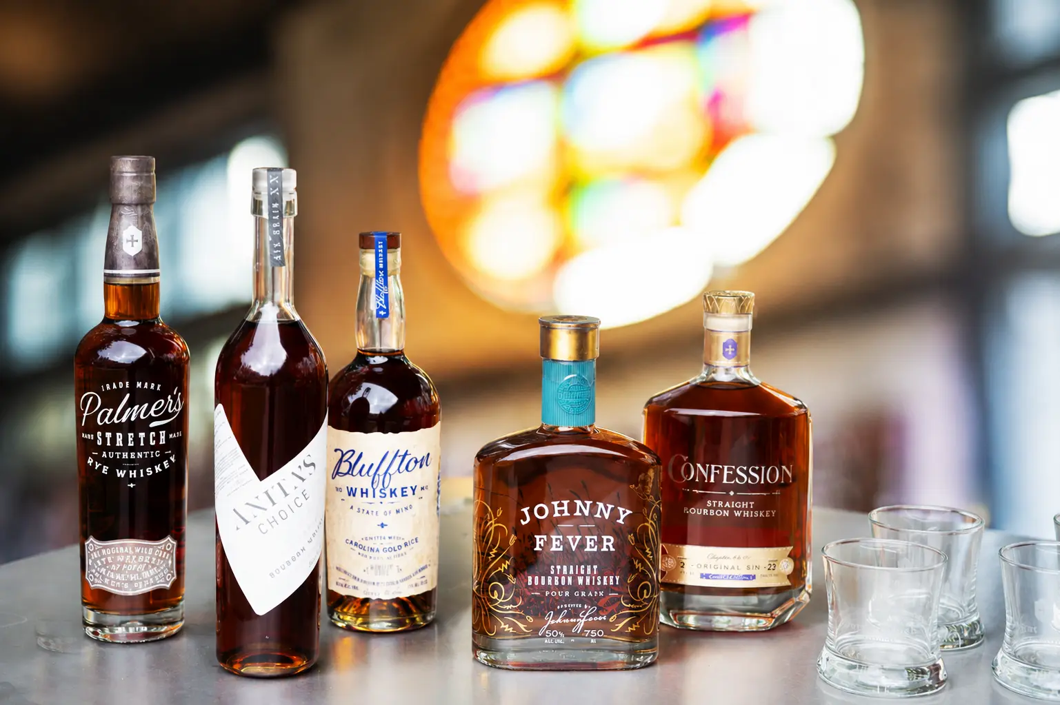 Five Burnt Church Distillery bottles — Palmer's Stretch Rye, Vanity's Choice, Bluffton Whiskey, Johnny Fever, and Confession — with a stained glass window behind.