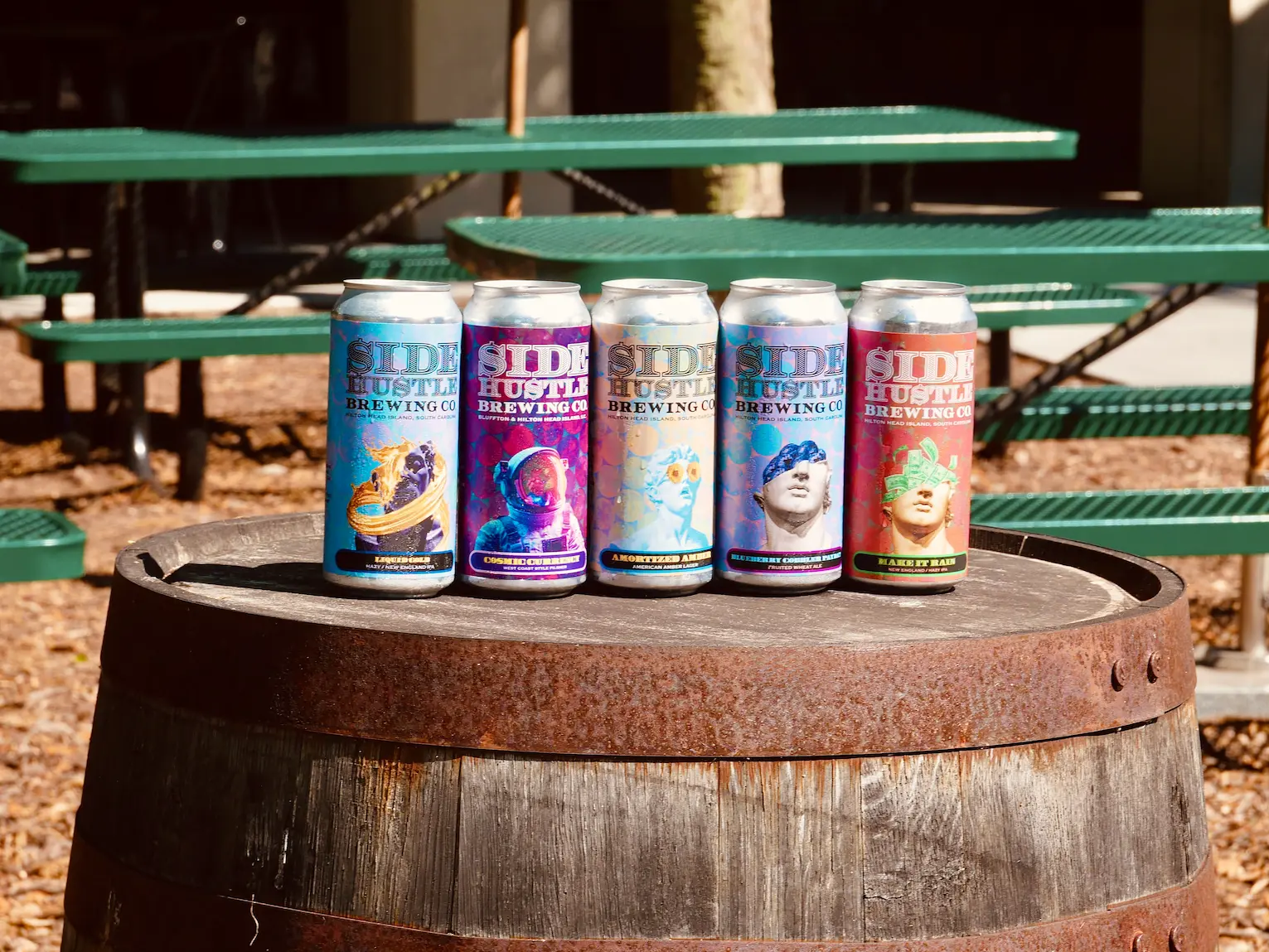 Five Side Hustle Brewing Co. cans lined up on a wooden barrel, with colorful classical-statue label art and picnic tables behind.